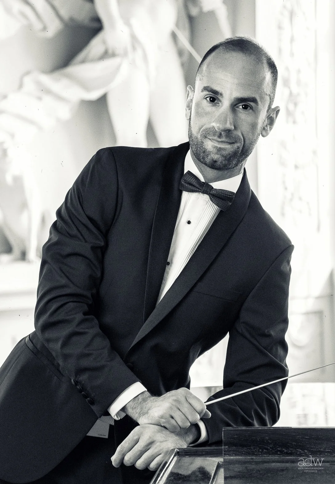 A man in a tuxedo with a bow tie, standing indoors near a window, holding a pointer, with a sculpture in the background.
