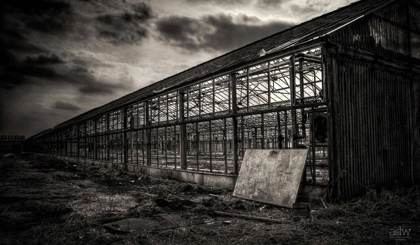 An abandoned, dilapidated greenhouse structure with a rusted, weathered exterior and missing glass panes, set against a dark, cloudy sky.