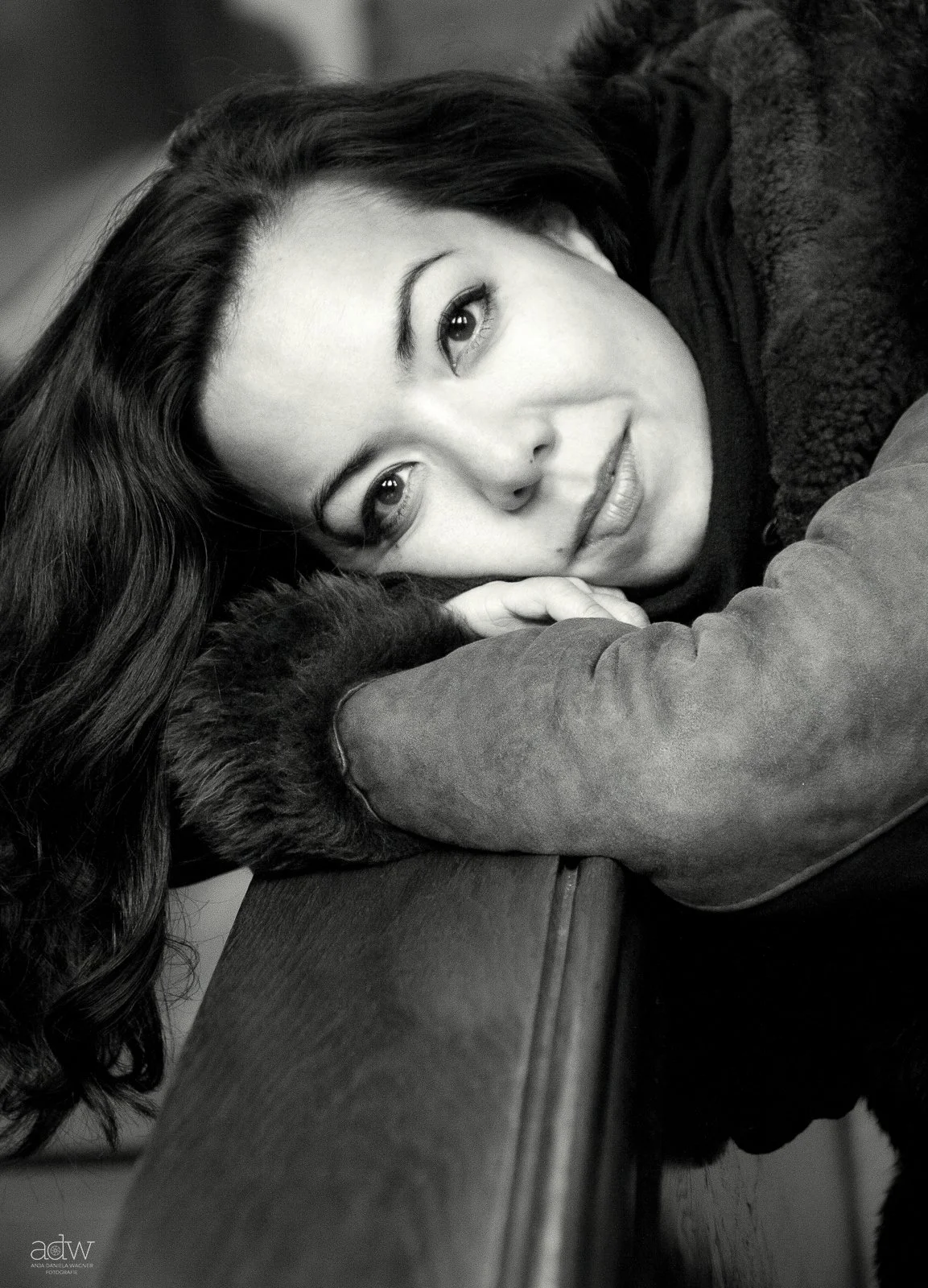 A black and white close-up photograph of a woman with long hair, lying down with her head resting on her arms and looking at the camera.