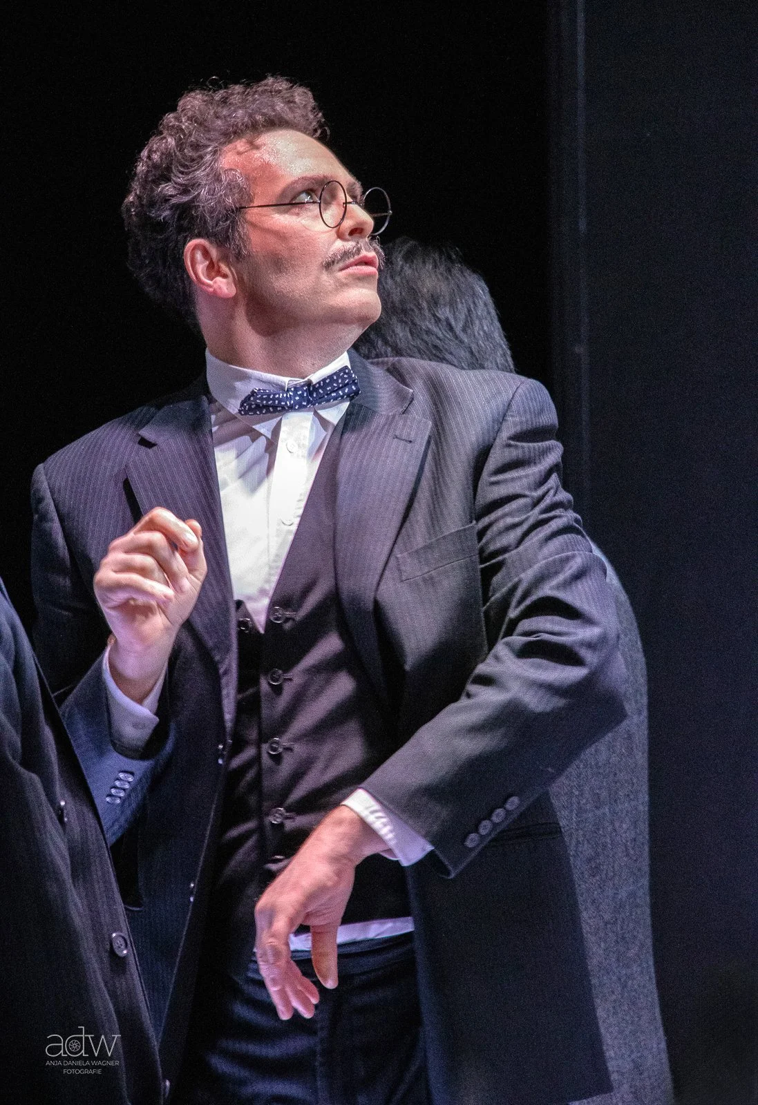 A man with curly hair, glasses, and a mustache wearing a pinstripe suit, white shirt, and blue bowtie, looking to the side with a thoughtful expression.