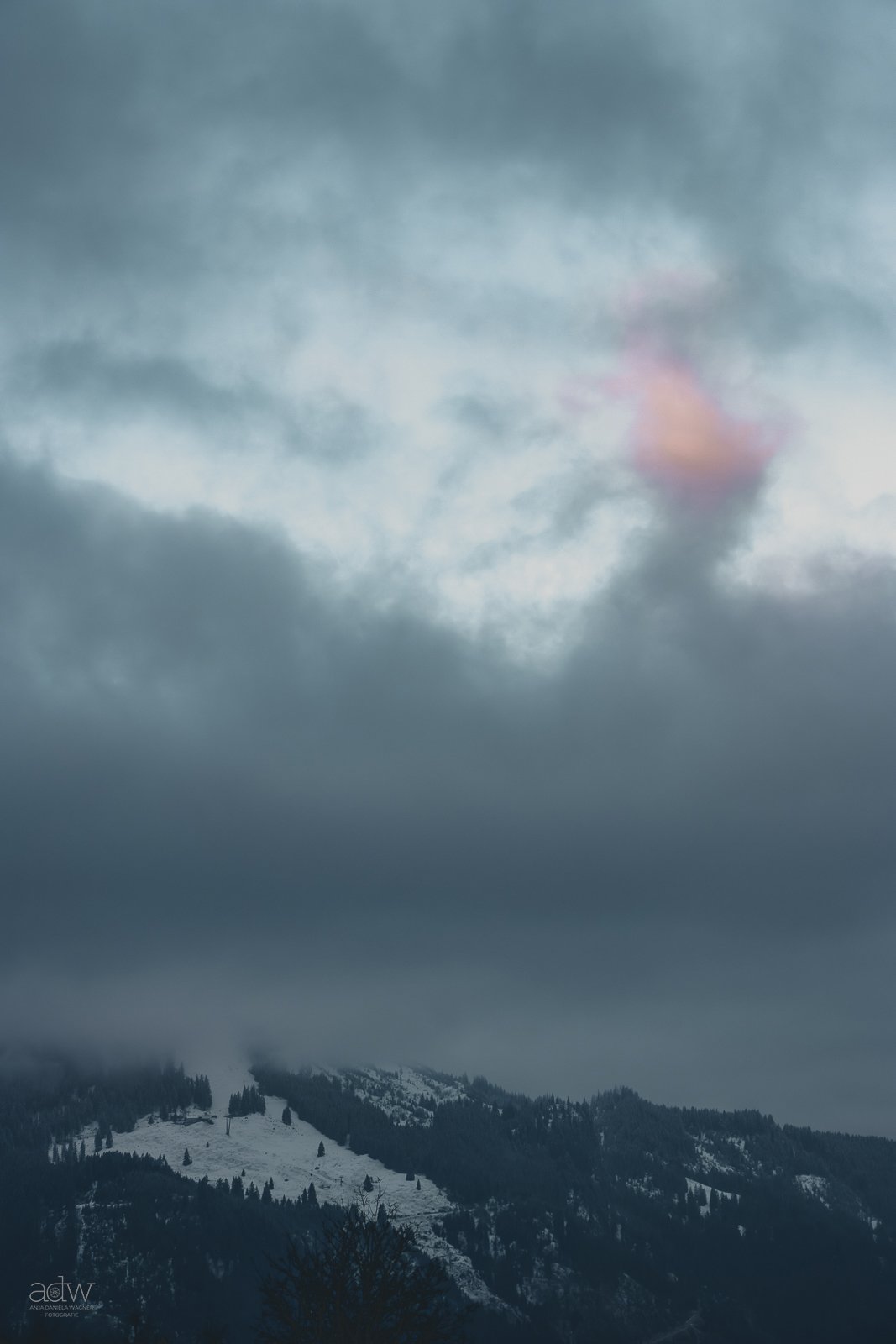 Cloudy mountain landscape with snow-covered slopes and dark forested areas, partly obscured by low-hanging clouds.