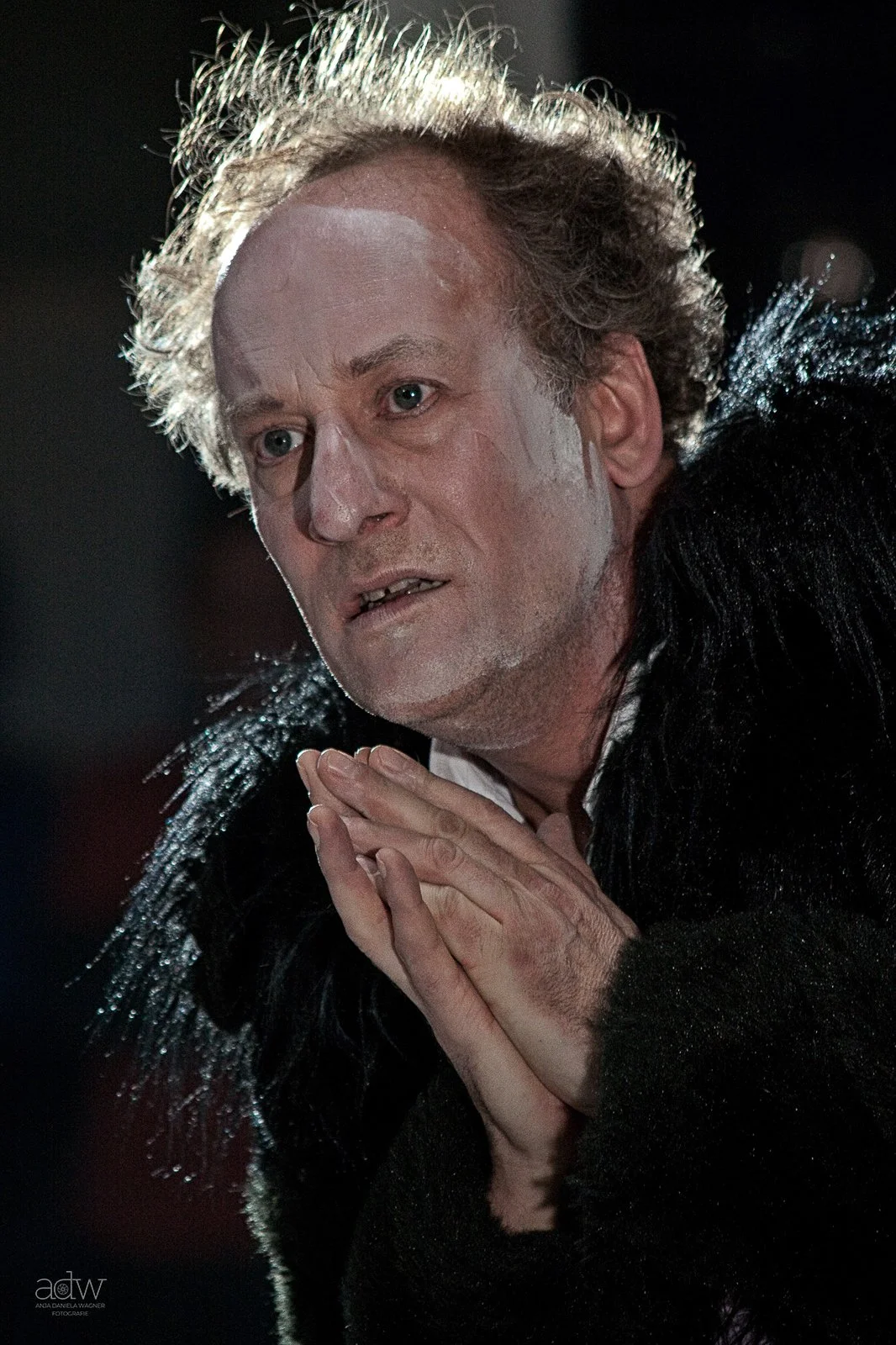 A man with curly, gray hair and light skin, wearing a black outfit with shiny black feather details, has makeup on his face and hands clasped in front of him, looking off to the side in a dark background.