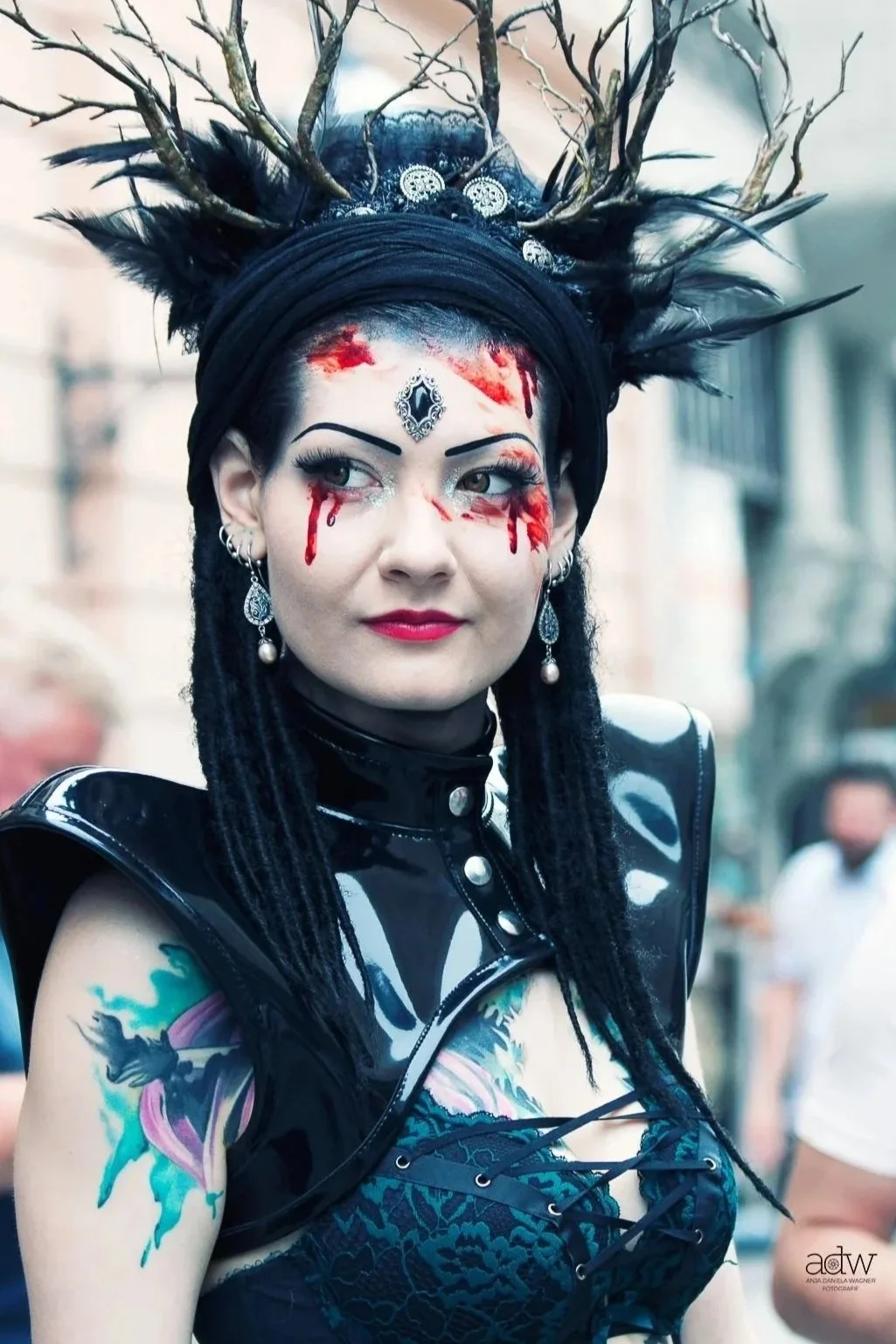 A woman dressed in gothic or fantasy cosplay with elaborate makeup, including red fake blood tears and silver forehead embellishments. She wears a black shiny vest, dark earrings, and a large headdress made of black feathers, lace, and antler-like branches. She has long black dreadlocks and a colorful tattoo on her upper arm.