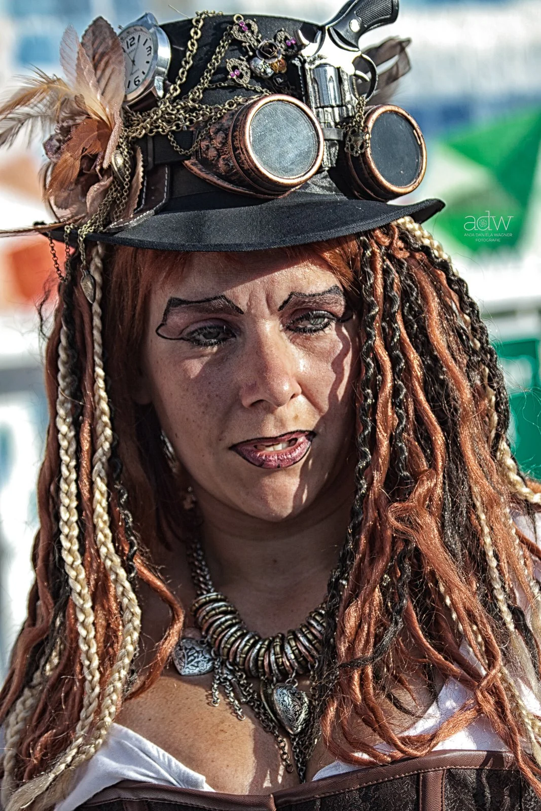 A woman with long, braided and curly hair wearing a black hat decorated with feathers, chains, a watch, glasses, and various jewelry, with makeup and a white top.
