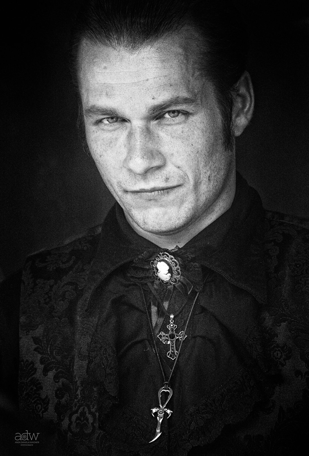 Black and white portrait of a man with slicked back hair, wearing a dark embroidered jacket with a brooch and multiple necklaces with crosses, looking directly at the camera.