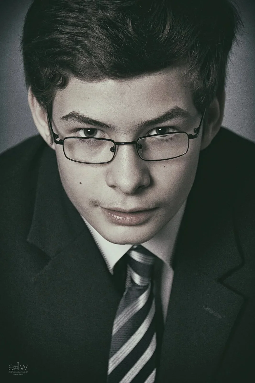 Close-up of a young man with glasses, wearing a black suit and striped tie, looking intently at the camera.