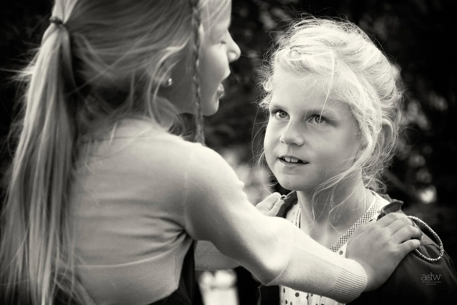 Two young girls facing each other, one with long hair tied back, the other with curly light hair, engaging in a conversation outdoors.
