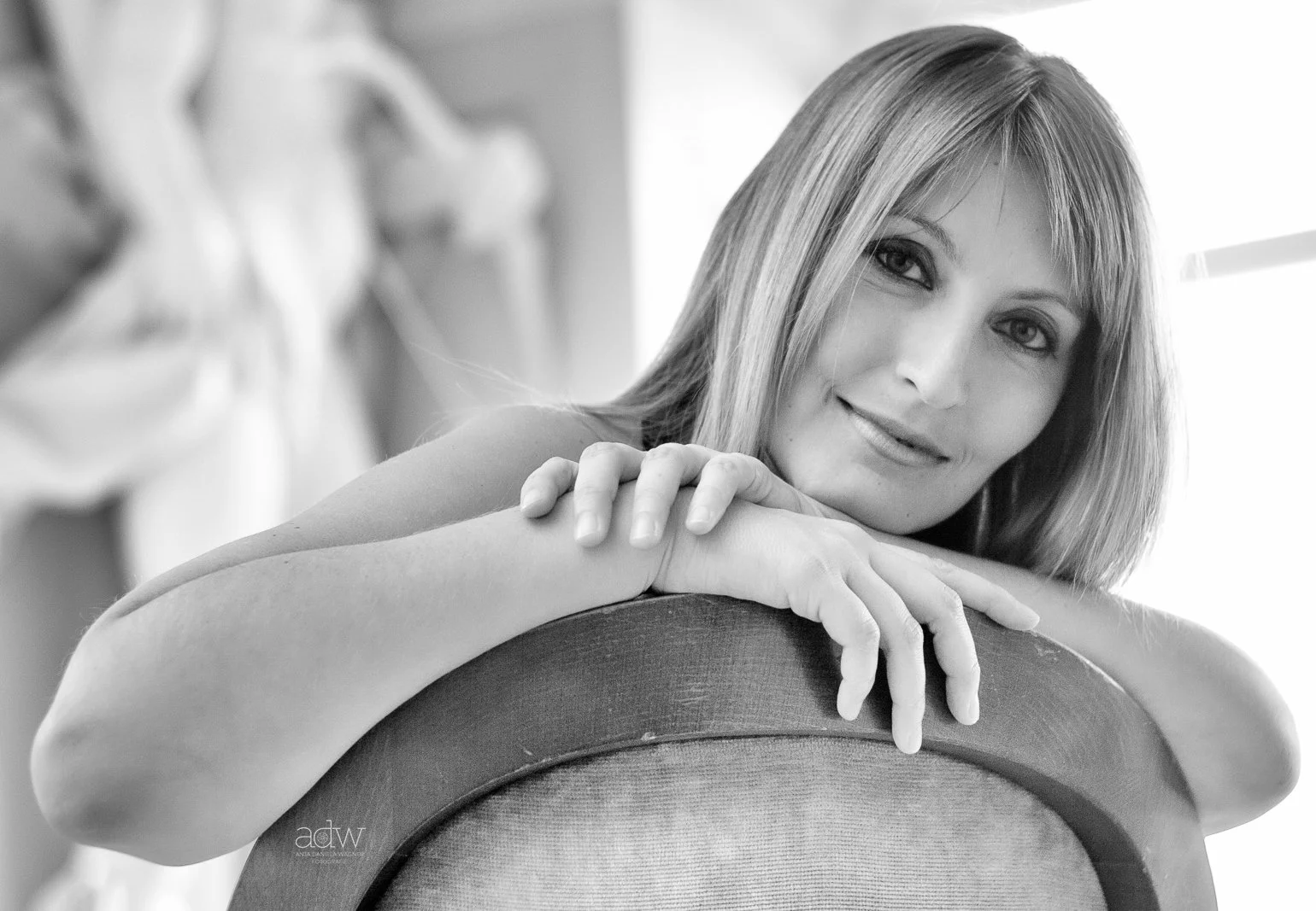 A woman with light-colored hair leaning on a chair, smiling softly, in a black and white photo.
