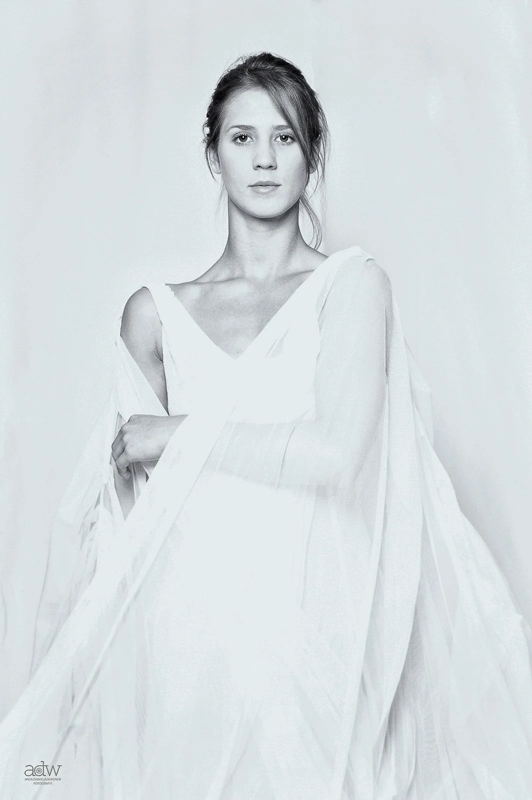 A woman with styled hair, wearing a white dress, stands against a plain background.