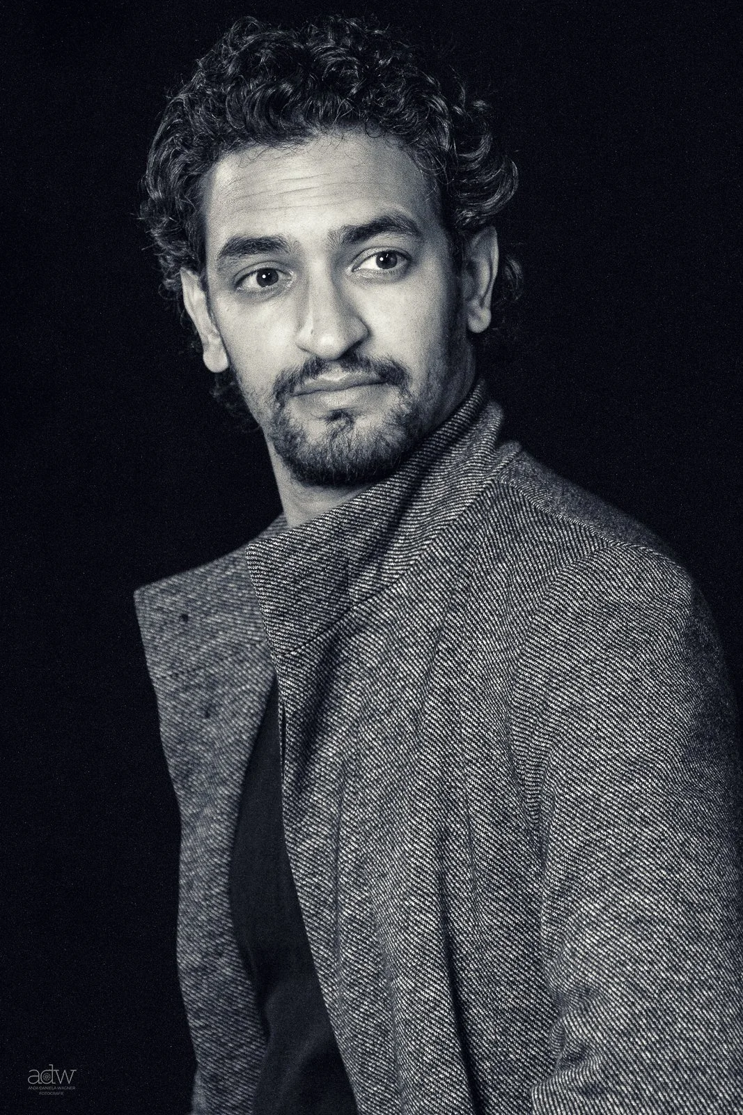A black and white portrait of a man with curly hair and a beard, wearing a textured blazer over a dark shirt, looking slightly to the side against a black background.
