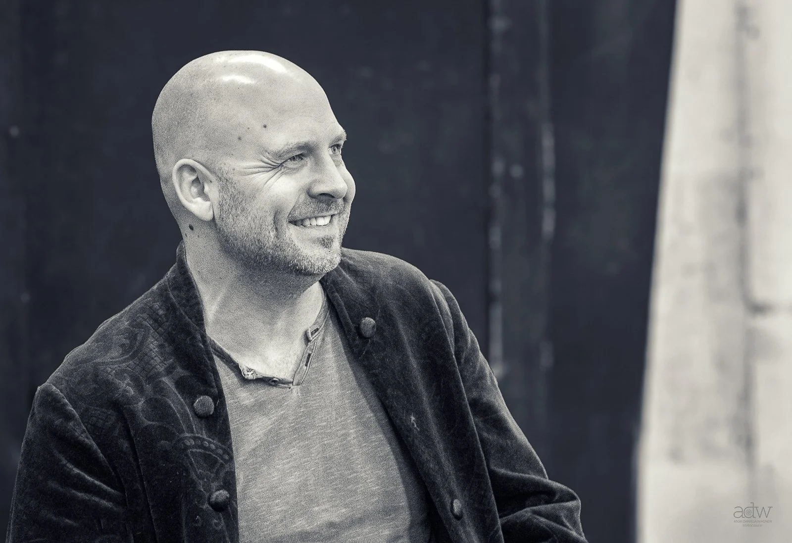 A smiling, bald man with a light beard wearing a dark jacket and a light-colored shirt, sitting indoors against a dark background.