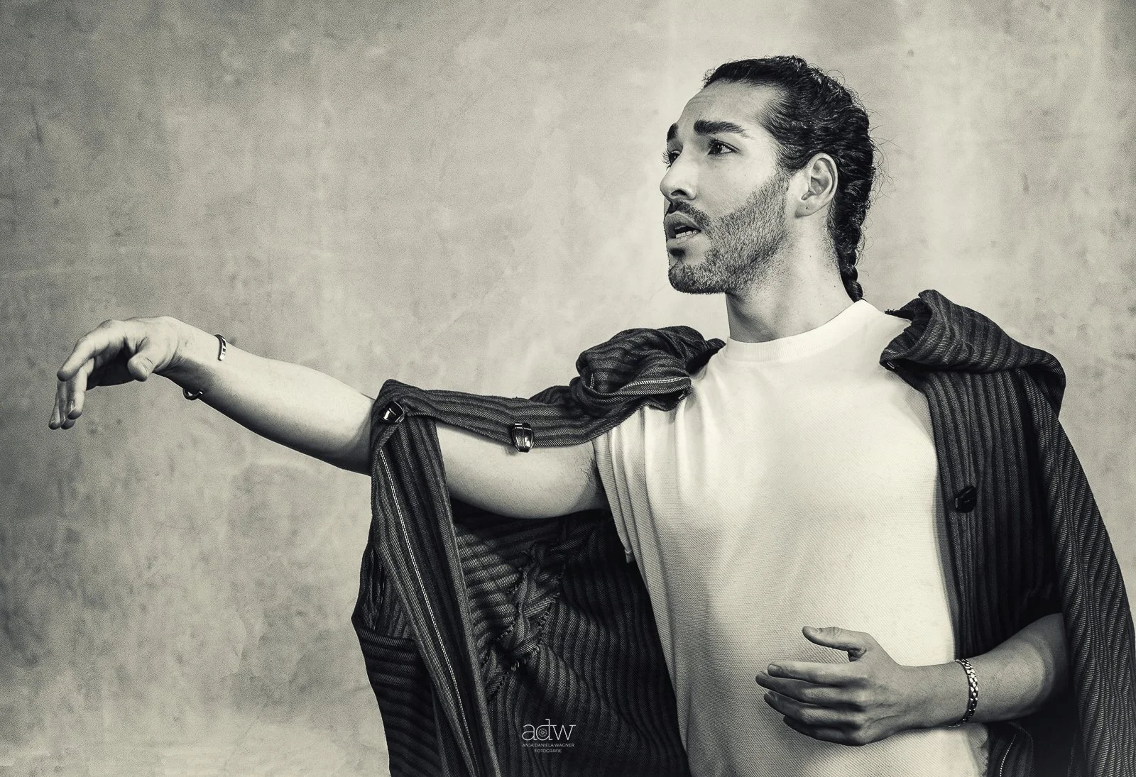 A man with a beard and slicked-back hair is wearing a white t-shirt and a striped jacket draped over his shoulders. He has an arm extended outward and is pointing or reaching, with a contemplative expression. The background is plain and textured.