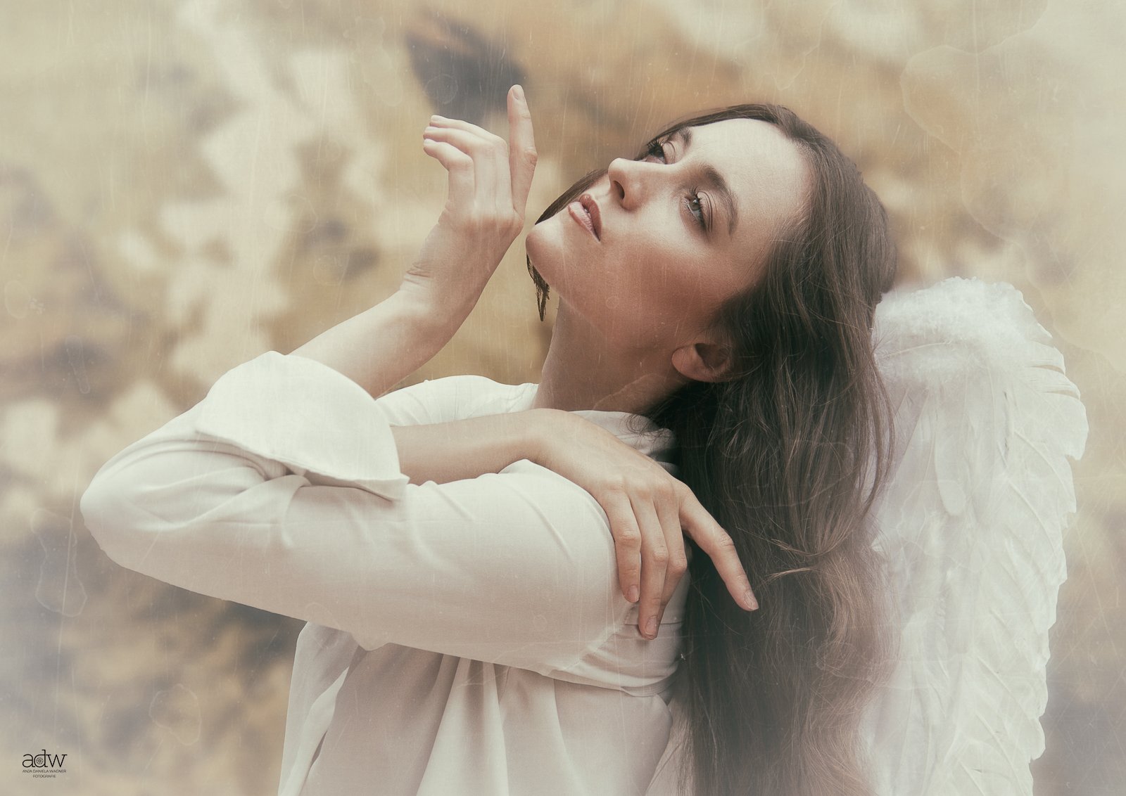 A woman with long brown hair, wearing a white dress and angel wings, is sitting in a relaxed pose with her eyes closed and head tilted back, touching her face gently with one hand in an outdoor setting with a blurred, warm-toned background.