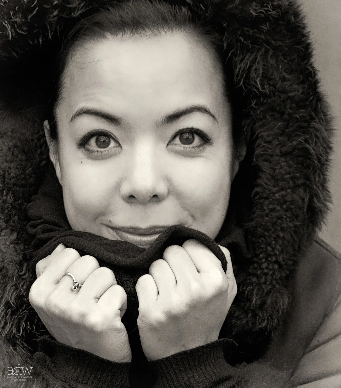 A woman with dark hair, light skin, and bright eyes, dressed warmly in a coat with a fur-lined hood and holding a scarf close to her face.