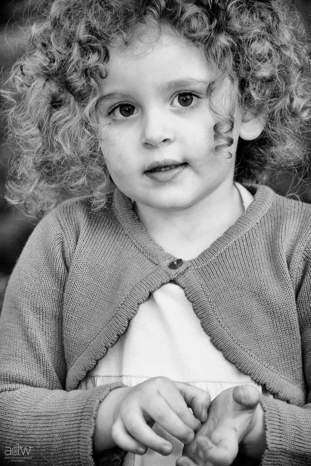 A young girl with curly hair wearing a cardigan and a dress, looking into the camera with her hands clasped.