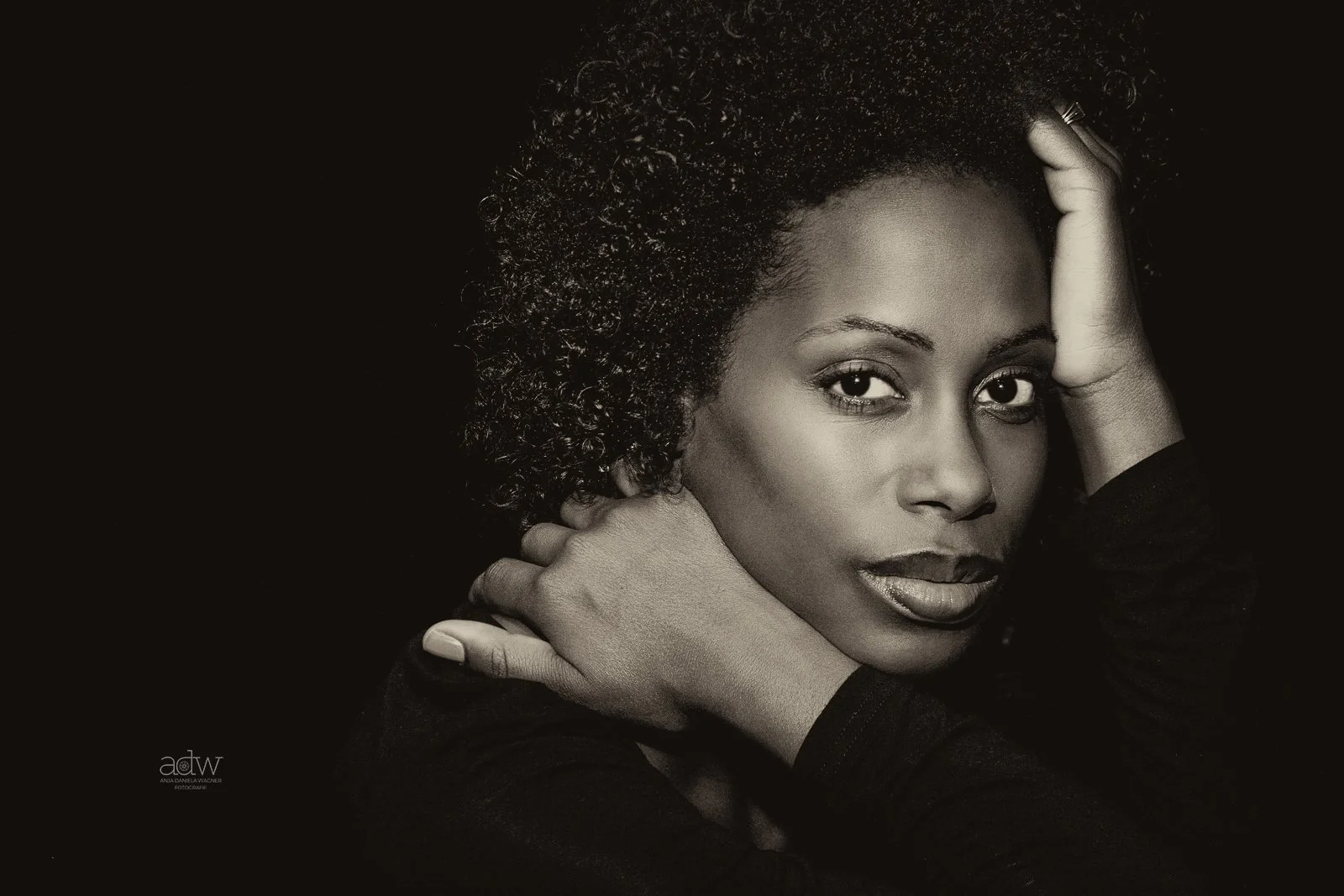 Black and white portrait of a woman with curly hair looking directly at the camera, hands raised near her face, against a dark background.
