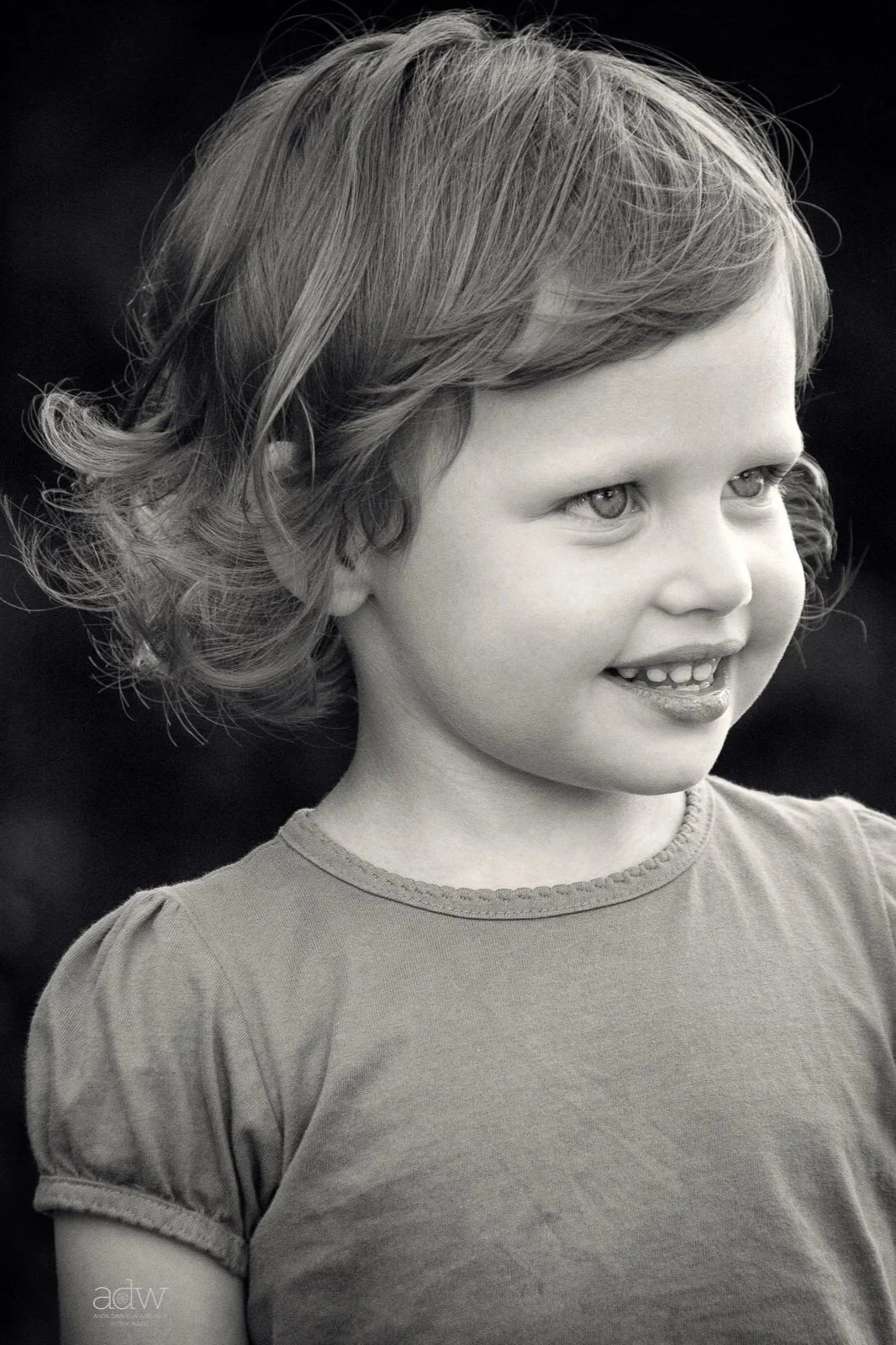 Close-up black and white portrait of young girl with curly hair, smiling and looking to the side, wearing a casual t-shirt.