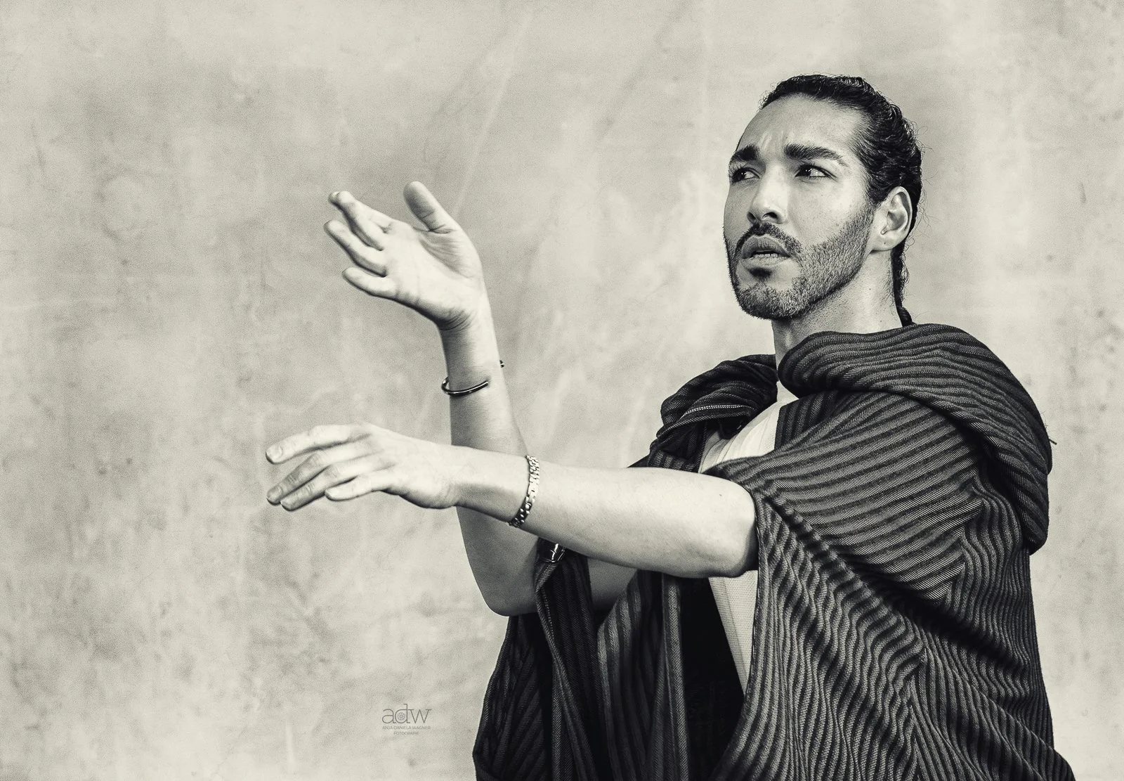 Black and white photo of a man with long dark hair, beard, and mustache, gesturing with his hands, wearing a striped cloak, against a plain background.