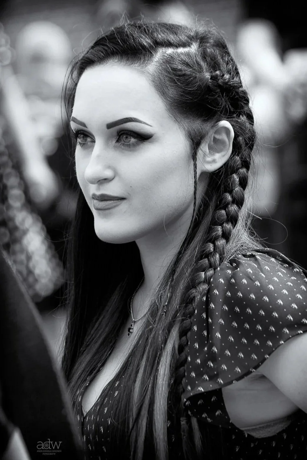 A black and white photograph of a woman with long hair styled into two French braids. She has makeup with defined eyebrows and eyeliner, and is wearing a patterned top and a necklace.