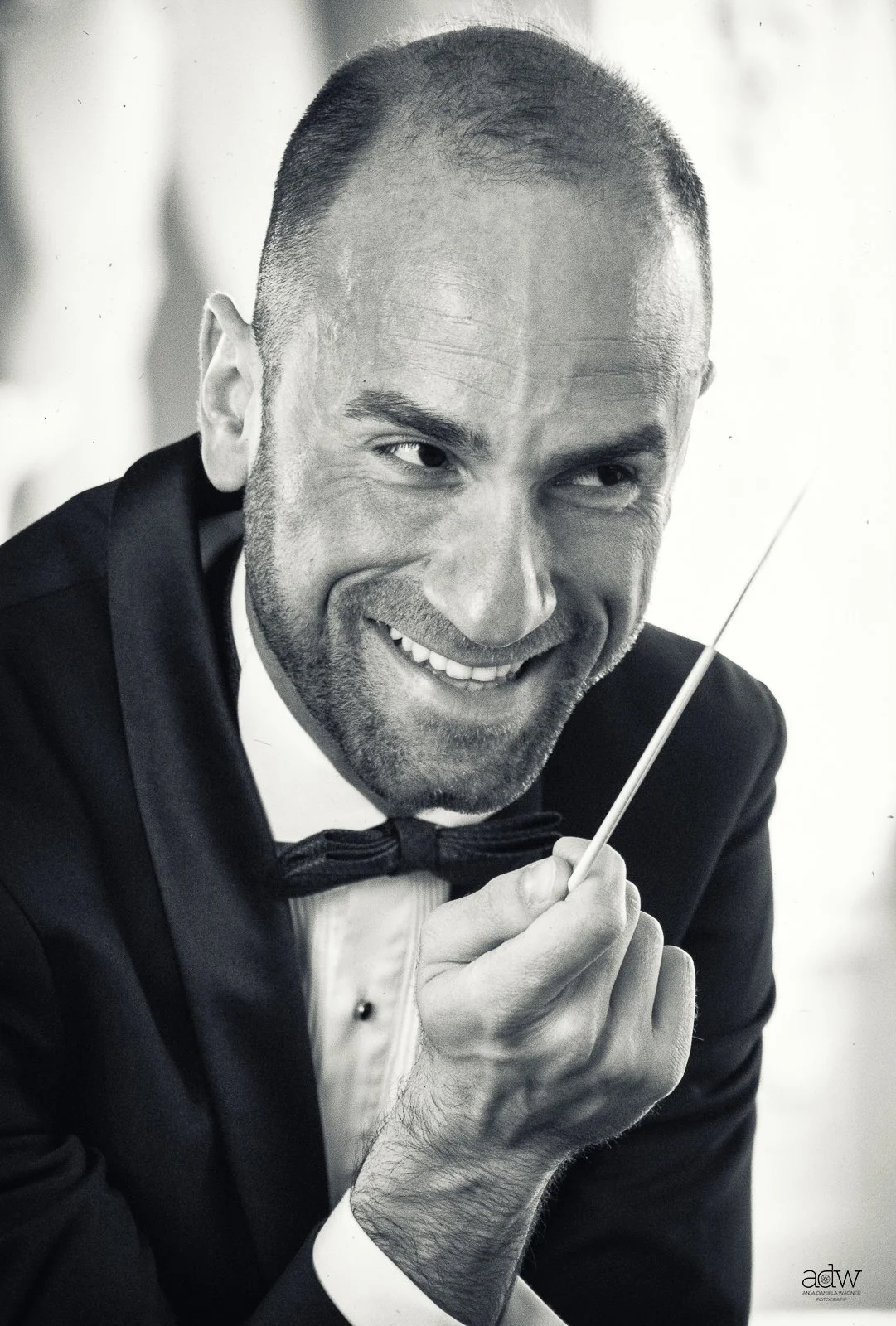 A man with a shaved head and beard, dressed in a tuxedo with a bow tie, smiling and holding a conductor's baton.