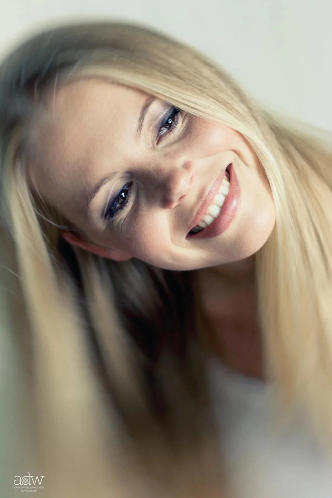 Close-up of a woman with long blonde hair smiling and tilting her head, with soft lighting and a neutral background.