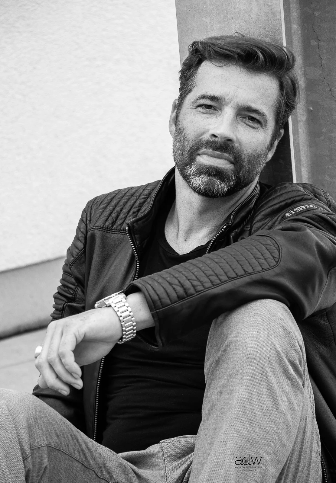 Black and white photo of a man with a beard, sitting outdoors, wearing a leather jacket, black shirt, and light-colored pants, with a wristwatch. He is leaning against a wall and looking at the camera.
