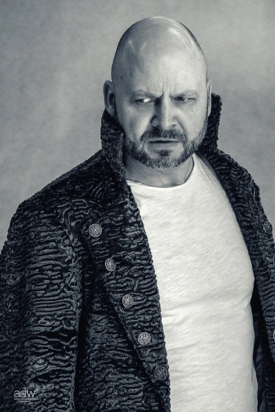 Black and white portrait of a bald man with a beard, wearing a textured coat with buttons over a white T-shirt.