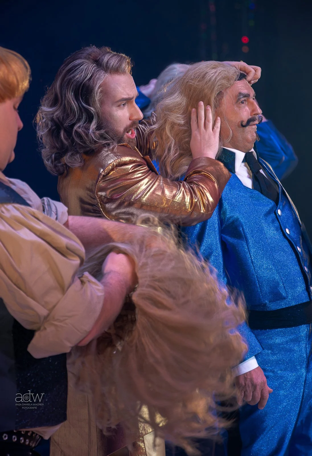 People dressed in colorful costumes, including a man with long gray hair and a mustache, and others adjusting wigs on a stage.