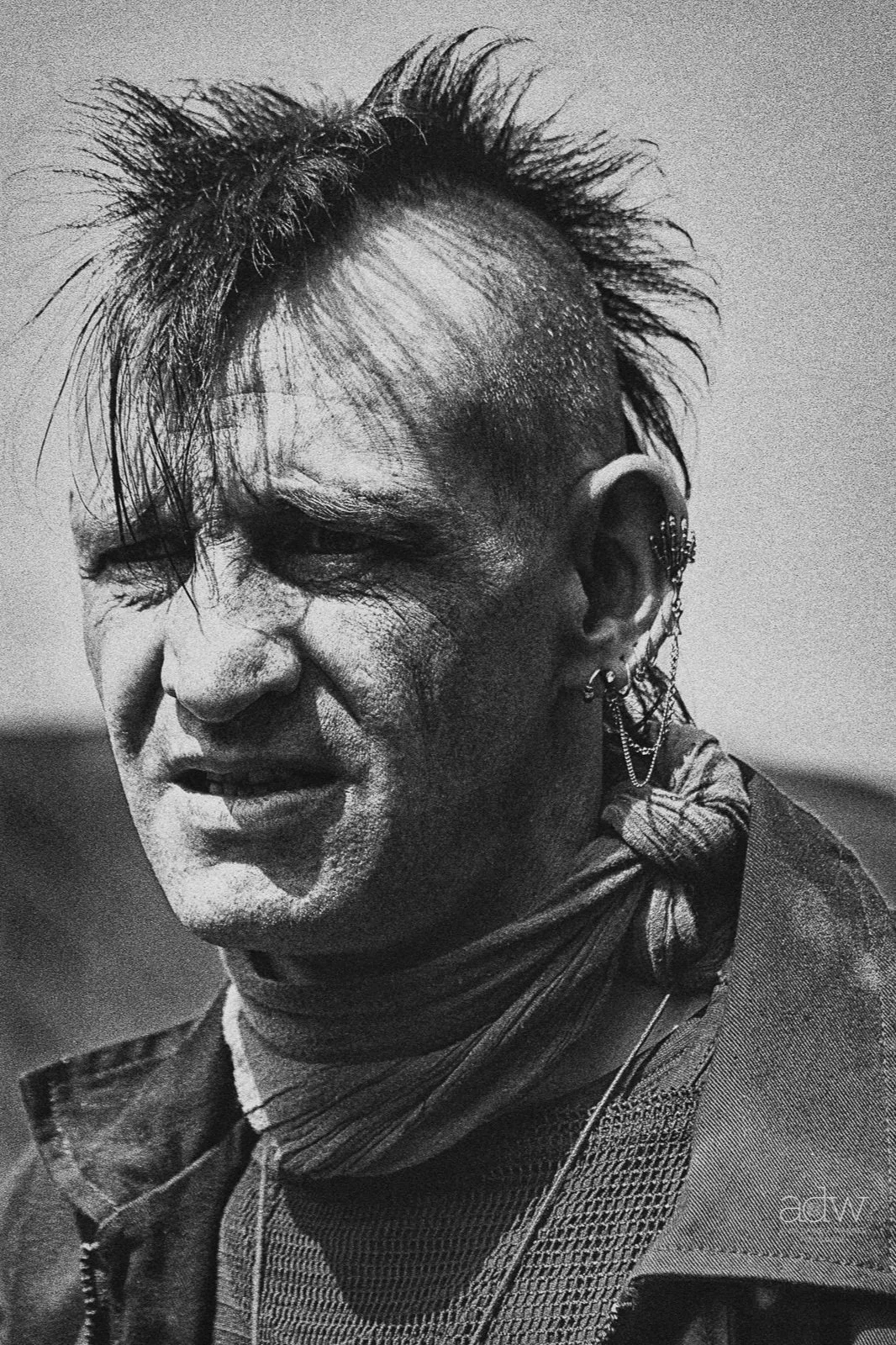 A black and white photo of a person with a punk hairstyle, multiple earrings, and layered clothing, making a grimacing face.