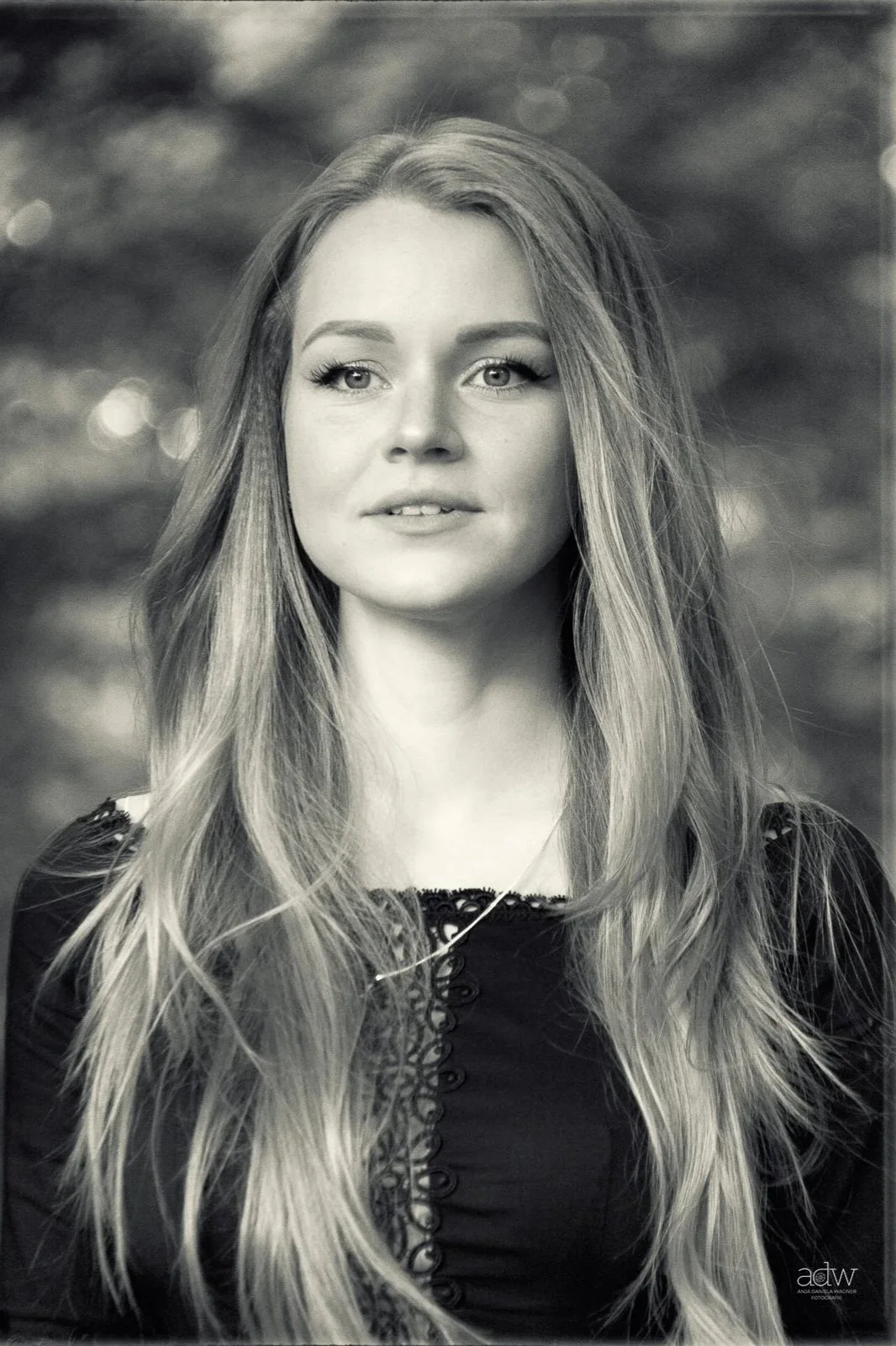 Black and white portrait of a young woman with long, wavy hair, looking slightly to the side. She has bright eyes, light makeup, and is wearing a dark top with decorative buttons and lacing detail. Blurred background with bokeh lights.
