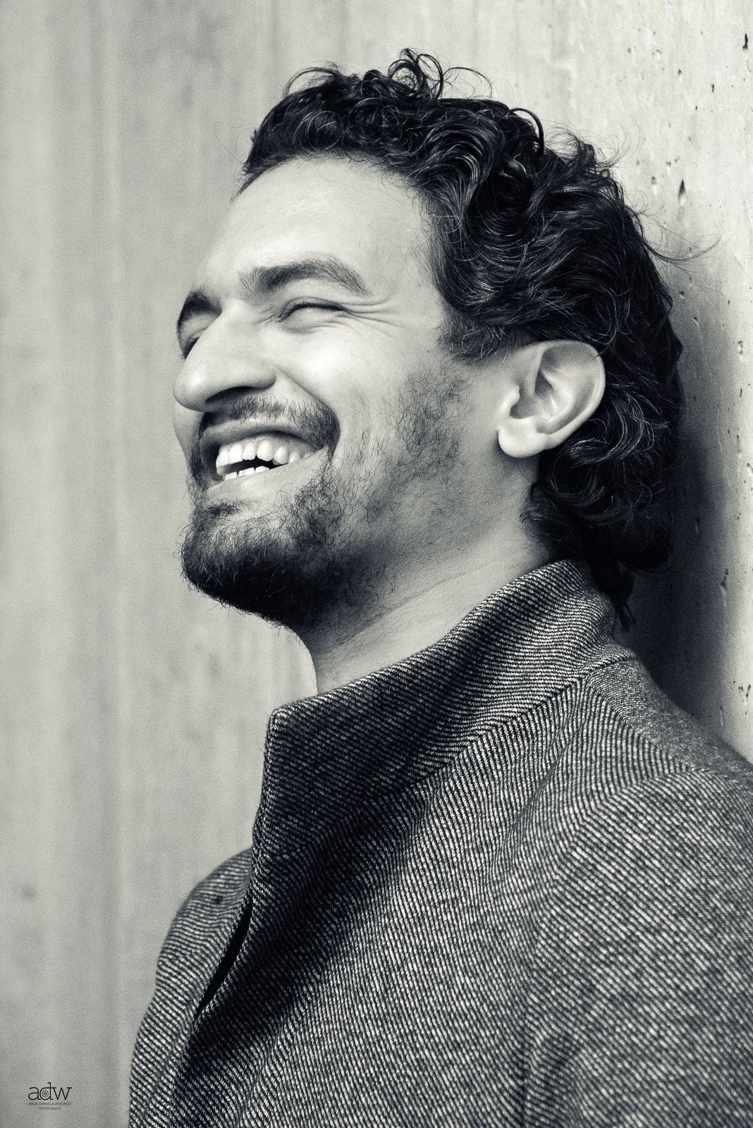 A black and white photo of a young man with curly hair and a beard, smiling and leaning against a wall.