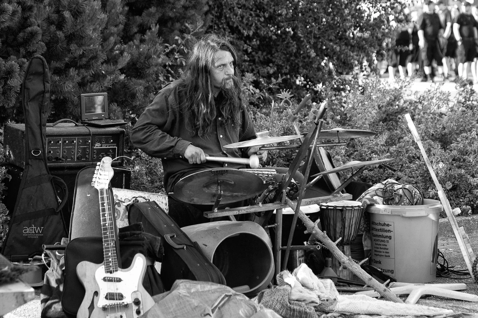 A street musician with long hair and a beard playing drums among various musical equipment and personal belongings, with a group of people walking in the background.