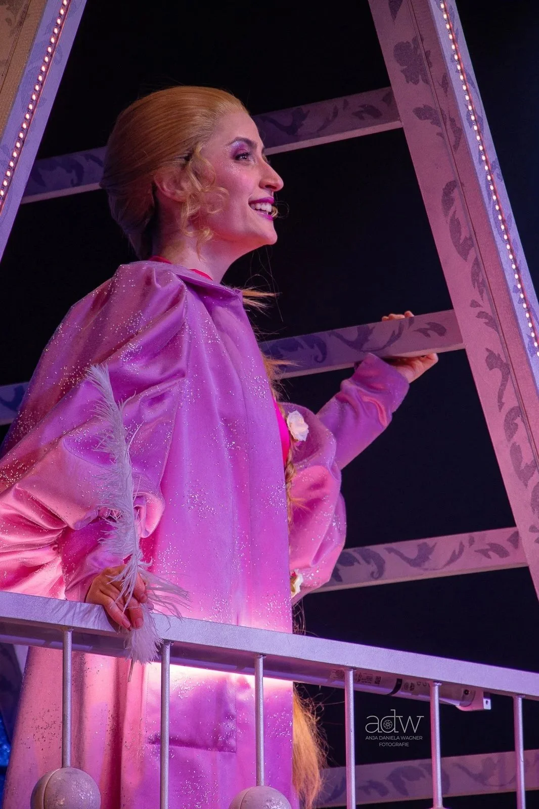 Side view of a woman dressed as a fairy tale princess wearing a pink shimmering dress with mirror details, standing next to a decorative purple arch, holding the arch with her right hand, and smiling.