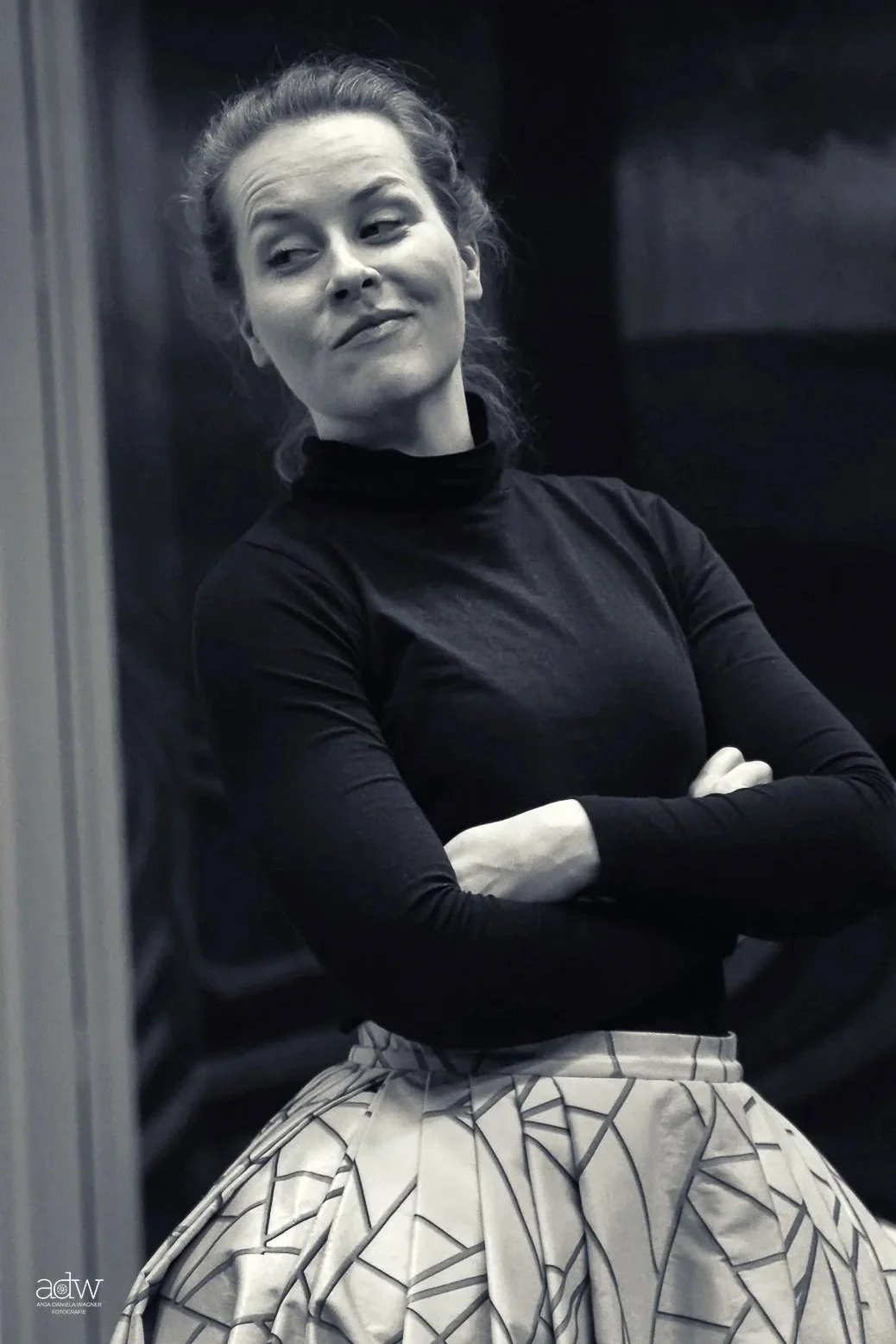 Black and white photograph of a woman with her arms crossed, wearing a long-sleeved turtleneck and a patterned skirt, standing indoors with a subtle smile and confident expression.