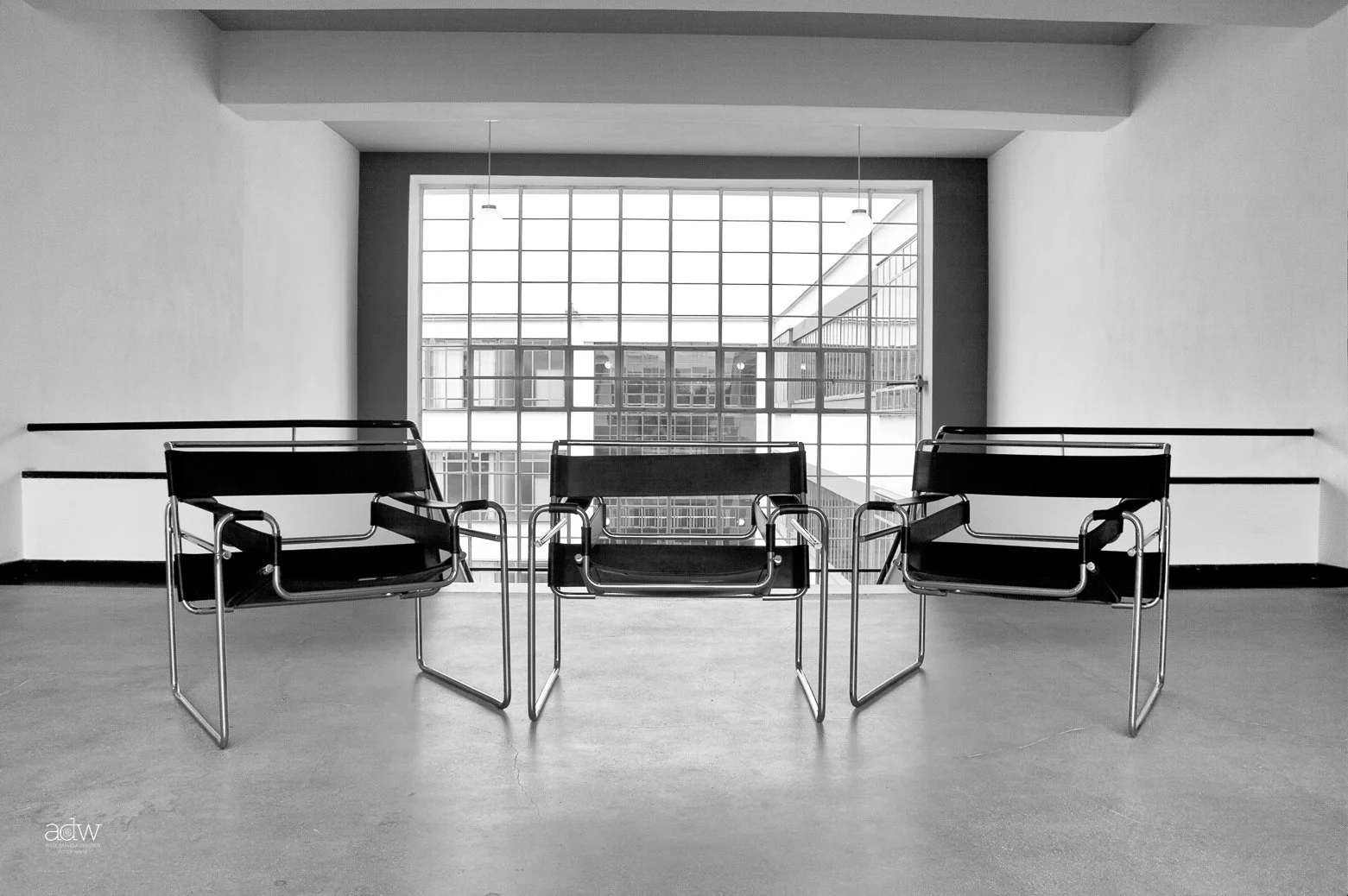 Three black chairs with metal frames placed in front of a large window with a grid pattern, in a minimalistic room with plain walls and a concrete floor.