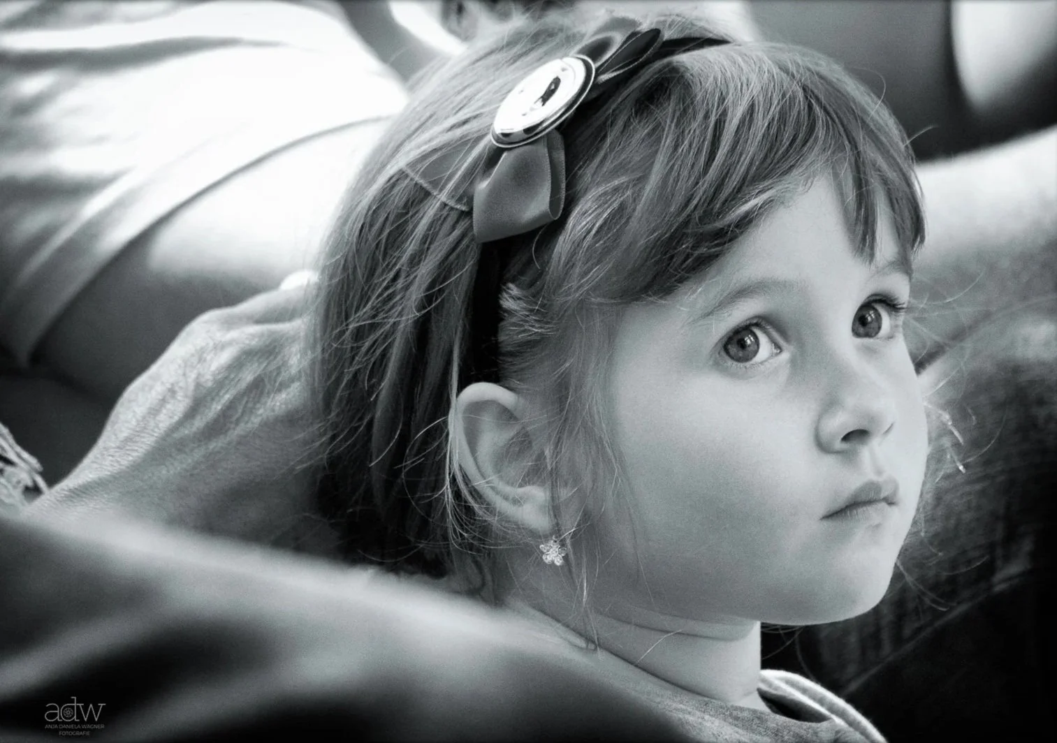 A young girl with light-colored hair wearing a bow headband and earrings, looking thoughtfully to the side, in black and white.