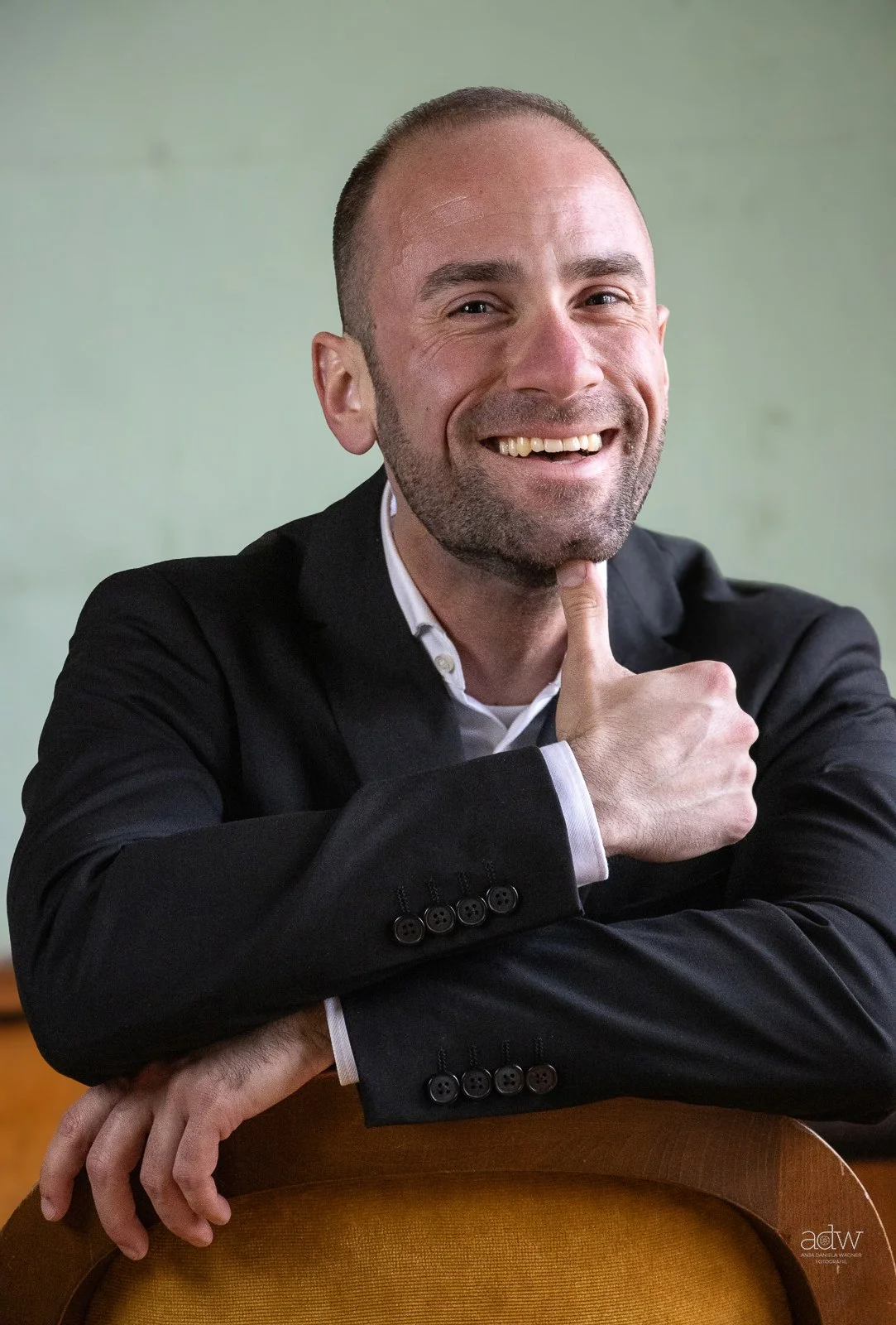 A smiling man in a black suit with a white shirt sits at a wooden table with a green background, resting his right arm on the chair in front of him, with his left hand touching his chin.