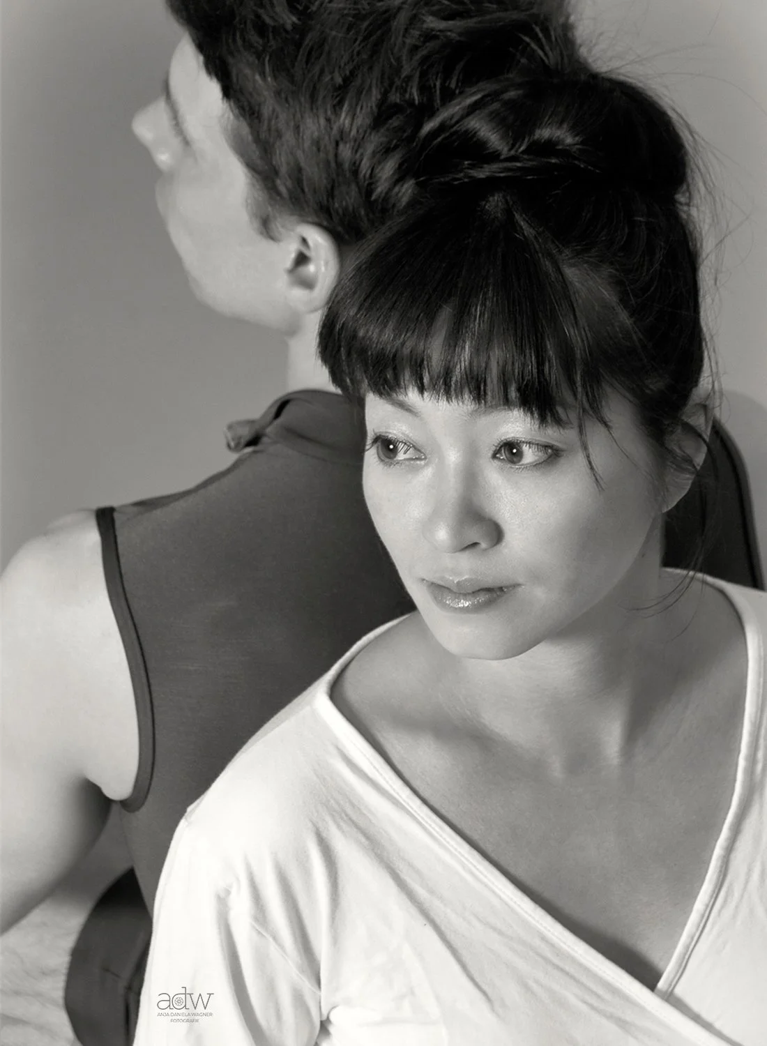 A black and white photo of a woman with short dark hair and bangs, looking to the side, with a man with short hair in the background facing away.