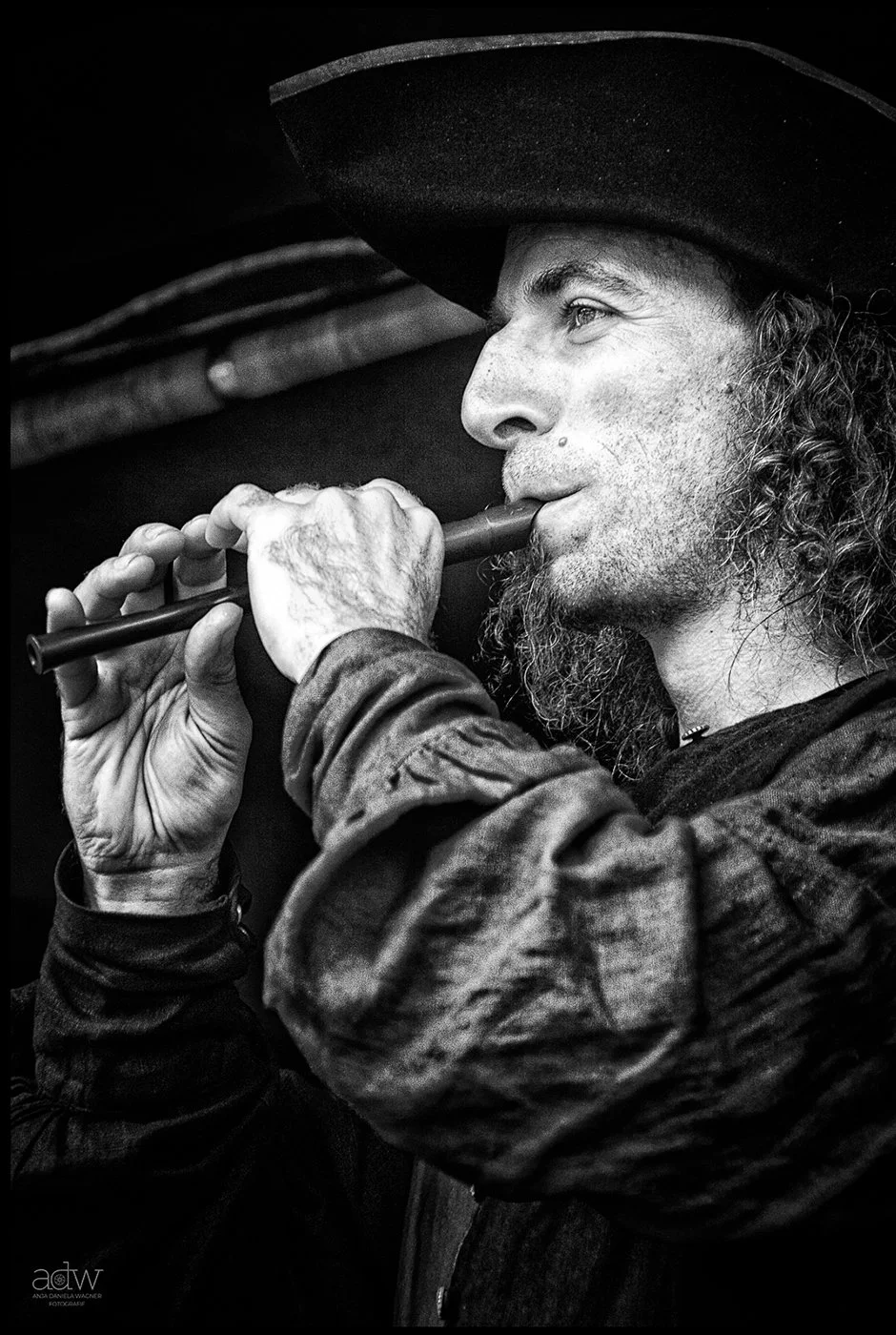 A black and white photo of a man with long curly hair and a beard, wearing a wide-brimmed hat, playing a flute.