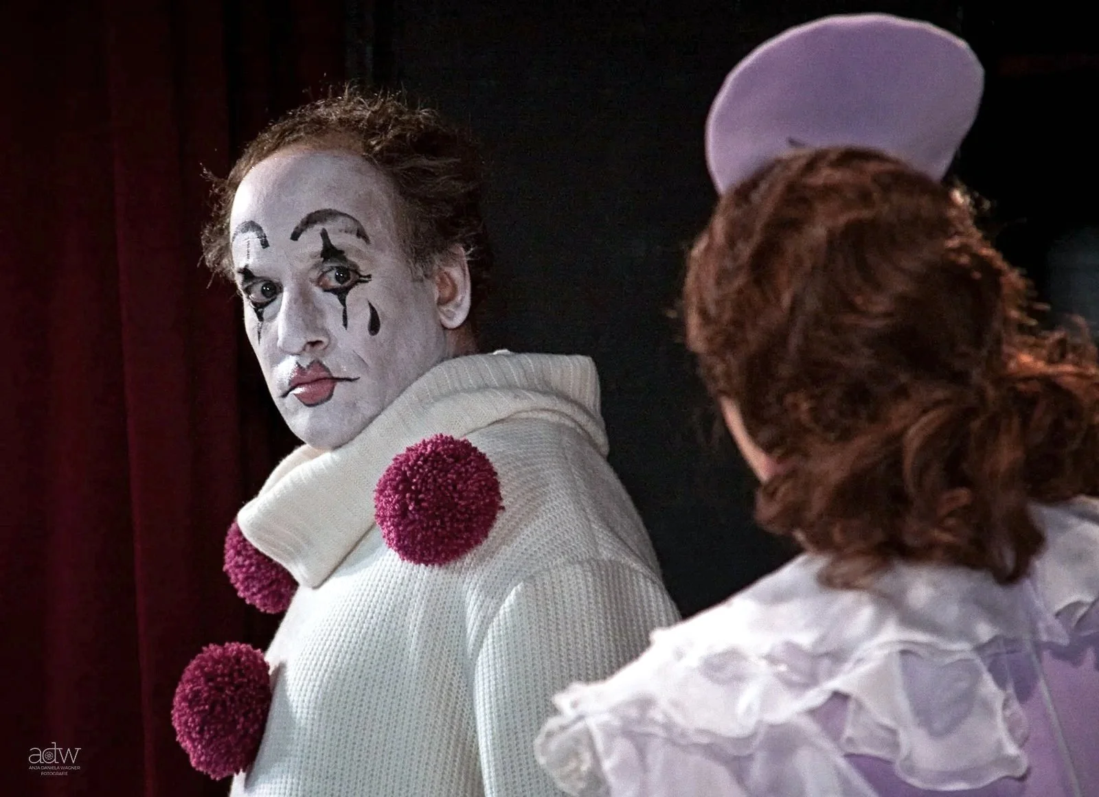 A person dressed as a clown with white face paint, black teardrops, and red lipstick, looking over their shoulder at another person with curly red hair and a purple hat.