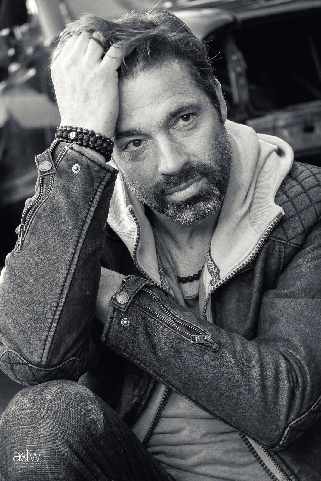 A man with a beard and tousled hair, wearing a leather jacket and layered clothing, rests his head on his hand, looking at the camera. He has bracelets on his wrist and is sitting in front of a blurred background.