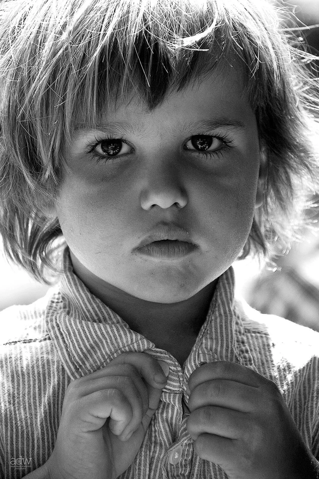 A young child with tousled hair looking directly into the camera, holding the collar of their striped shirt with both hands, in black and white.