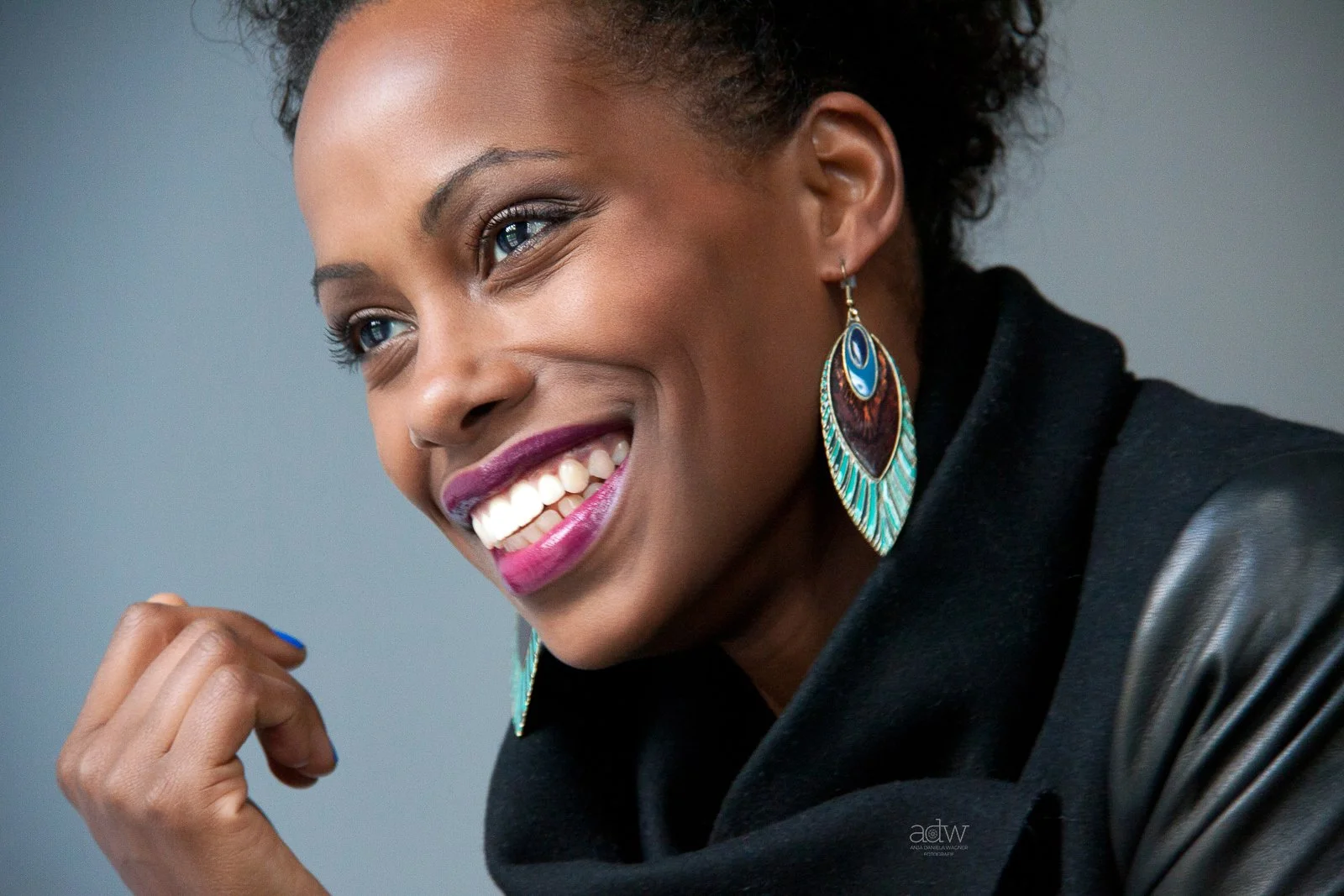 Close-up of a woman smiling with purple lipstick, wearing colorful earrings and a black top with leather sleeves.