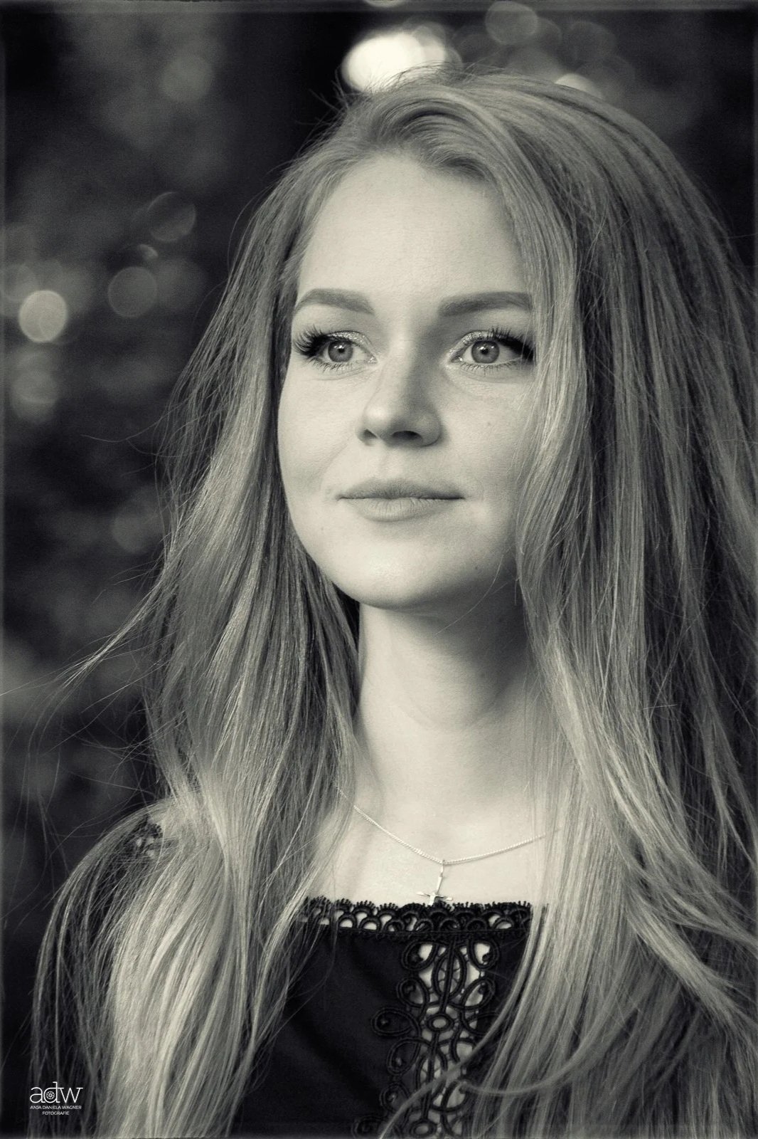Black and white portrait of a young woman with long, wavy hair, wearing a delicate necklace with a cross charm, and a lace detail on her top.
