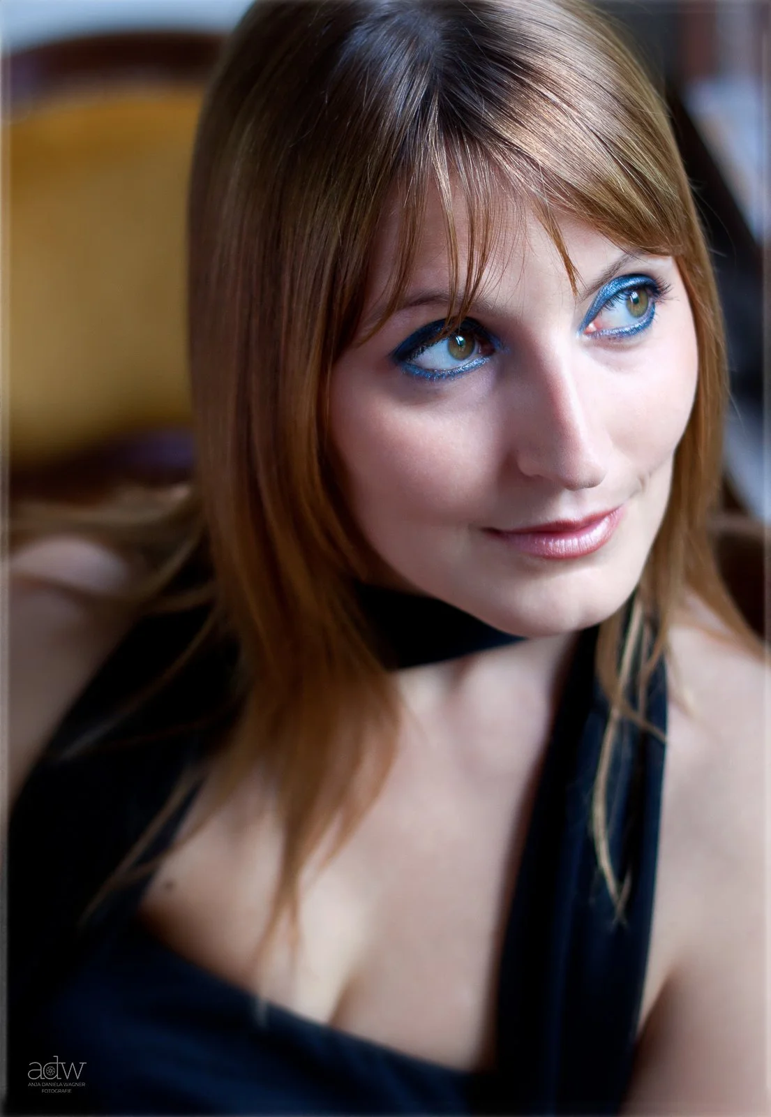 Close-up of a woman with reddish-brown hair and green eyes, wearing blue eyeshadow, smiling softly, with a blurred background.