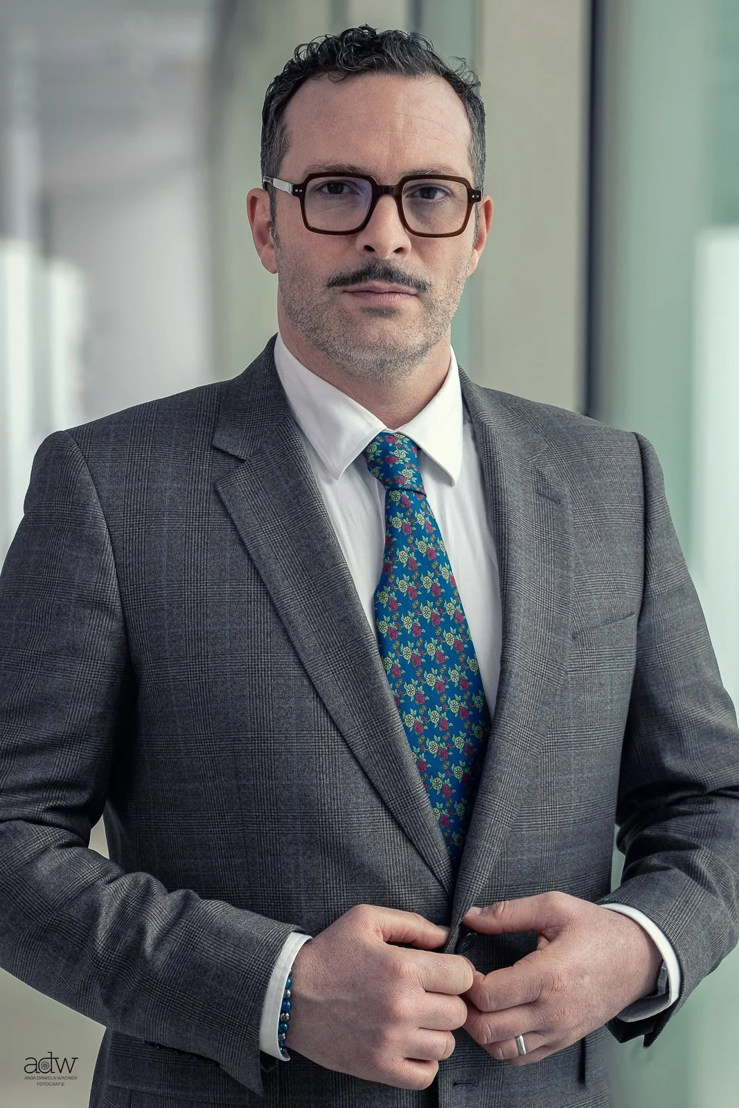 A man in a gray checkered suit, white shirt, and colorful patterned tie, wearing glasses, standing indoors with a blurred background of windows.