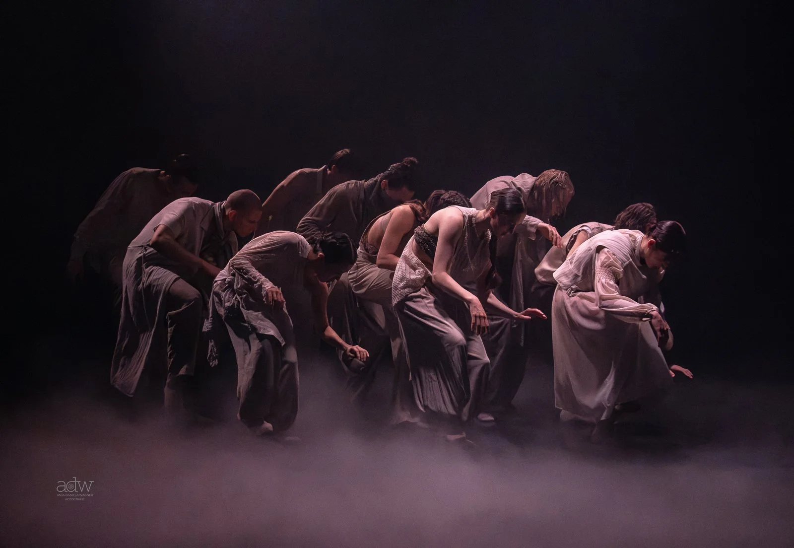 A group of dancers on stage, bowing with bent knees, dressed in loose, earthy-toned clothing, with a dark background and soft lighting