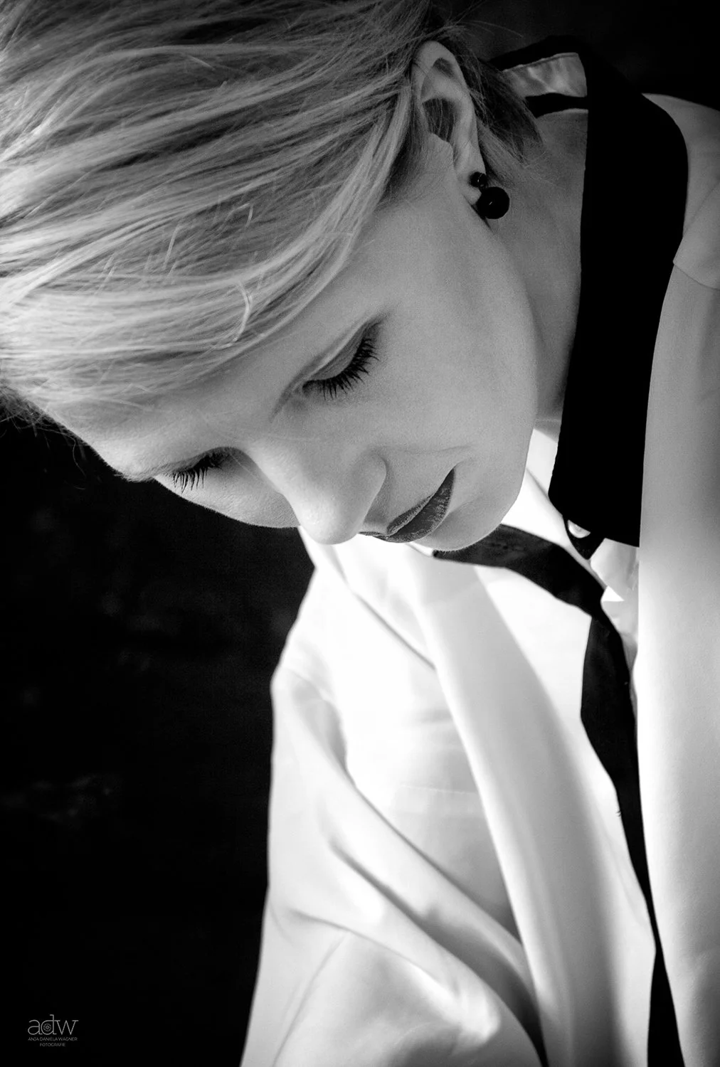 A close-up black and white photo of a woman with short blonde hair, wearing dark lipstick, and a white collared shirt with a black tie.
