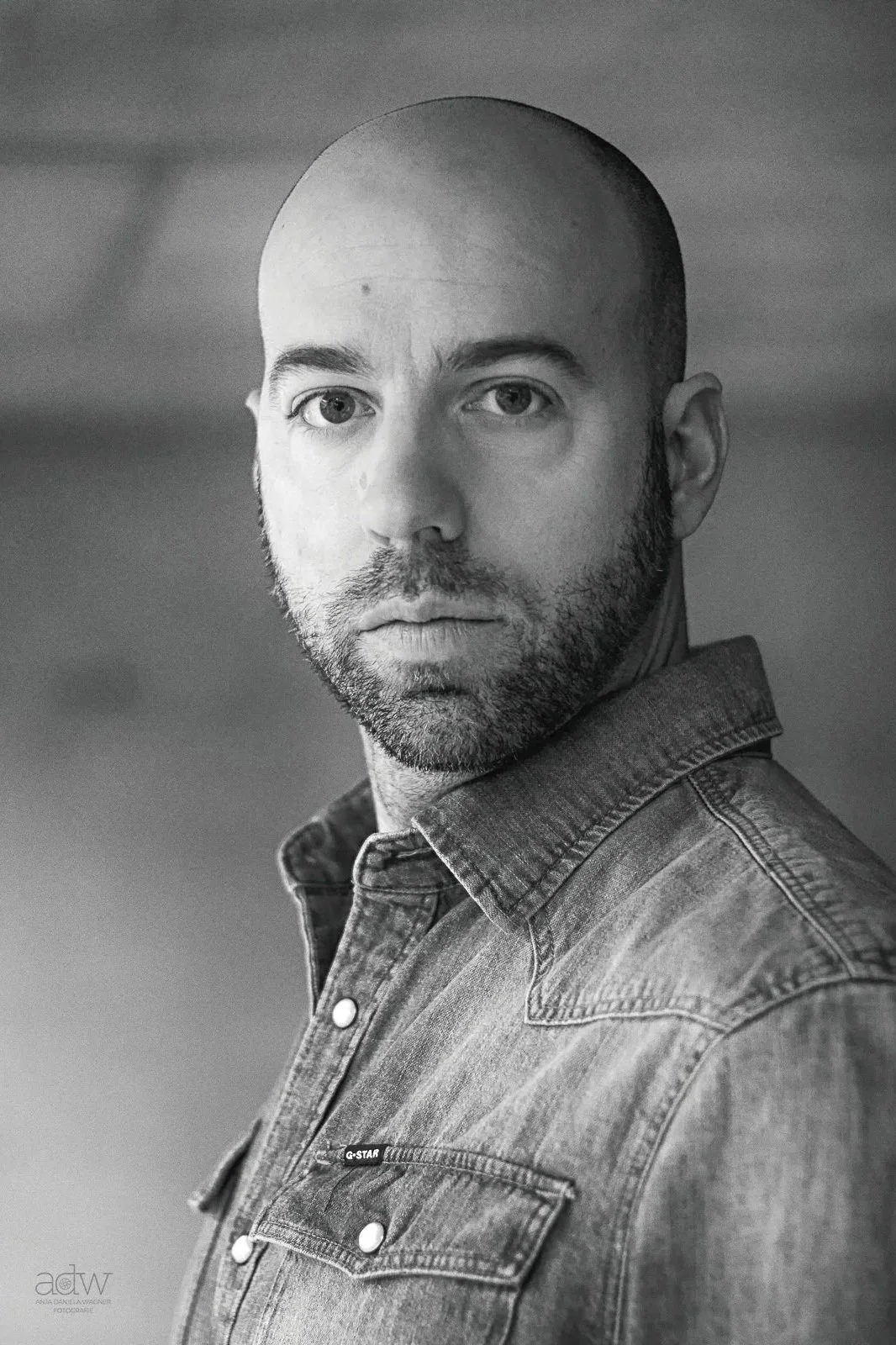 A black-and-white portrait of a man with a shaved head and a beard, wearing a denim shirt, looking directly at the camera.