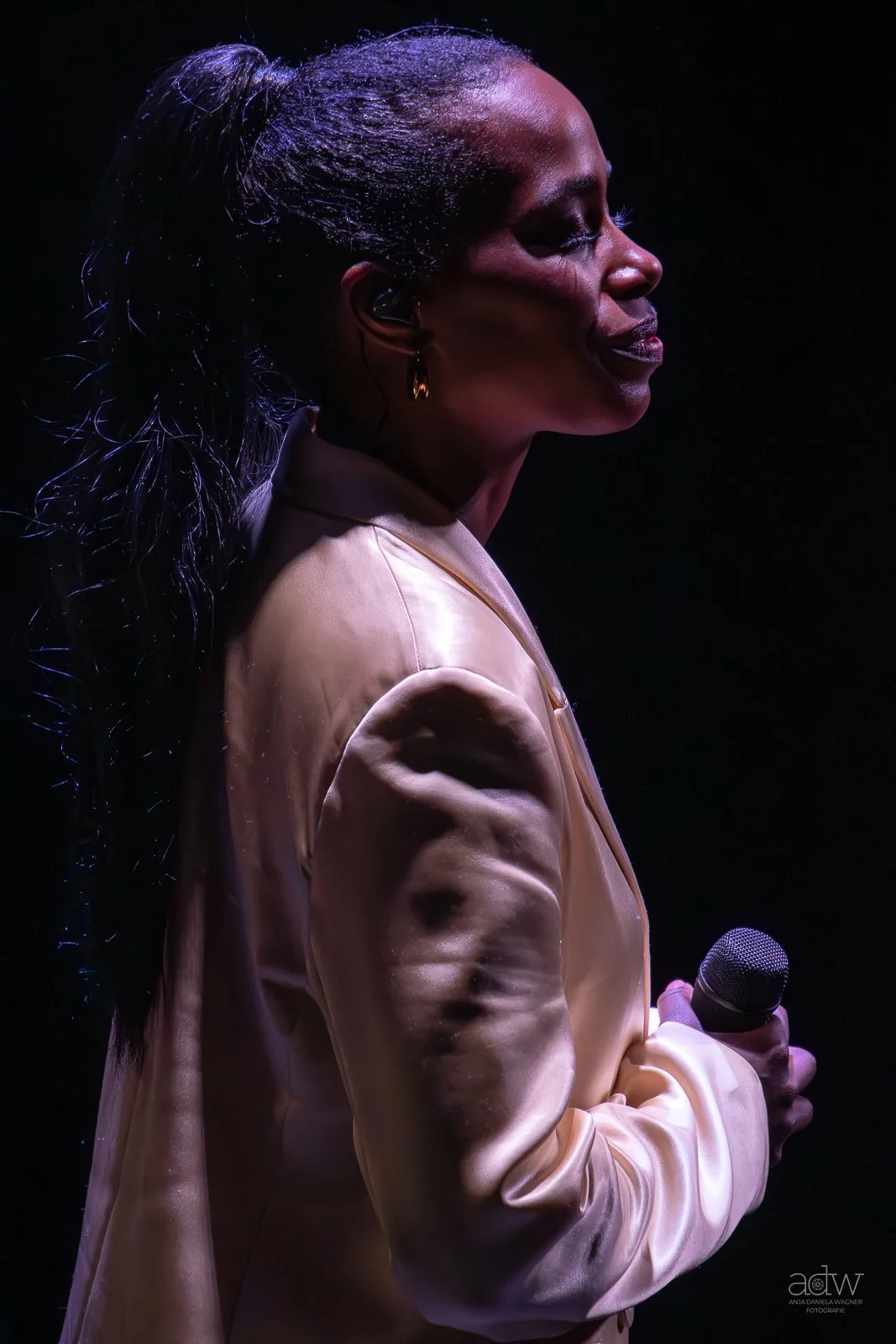 A woman singing on stage in a satin beige blazer, holding a microphone, with a dark background and dramatic lighting highlighting her profile.